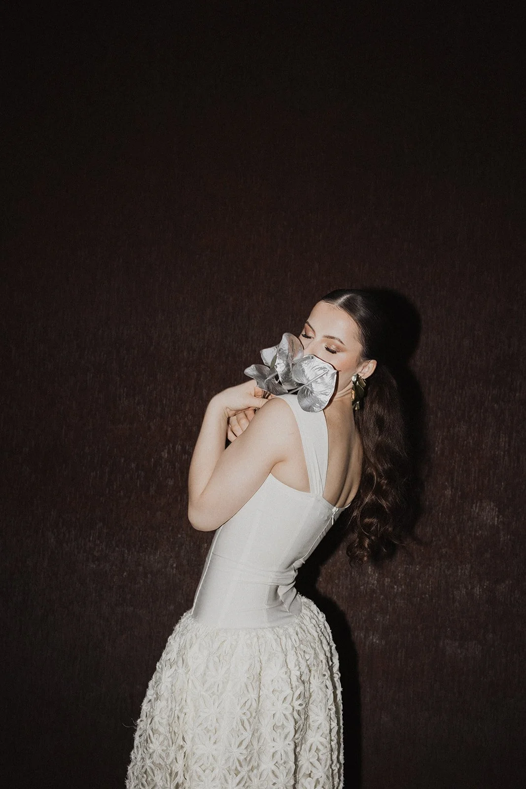Woman with dark hair in a white dress holding a metallic silver flower close to her face, standing against a dark textured background.