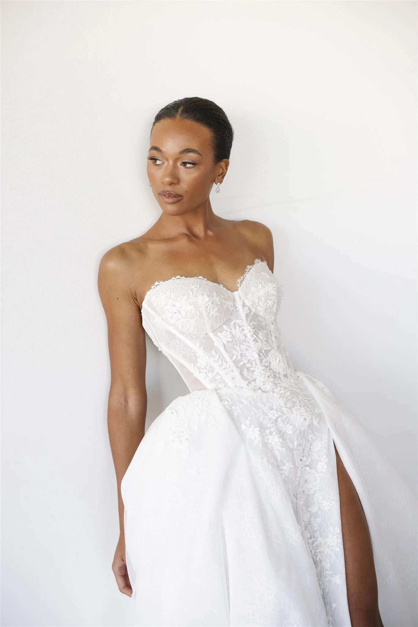A woman in a white wedding dress with lace details poses against a plain white wall.