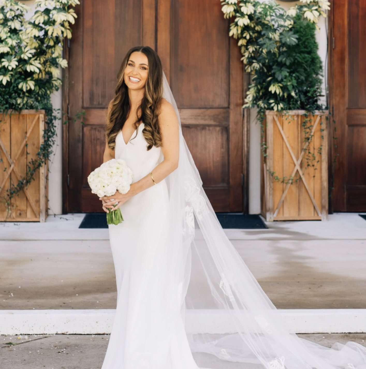 A smiling bride in a white wedding dress holding a bouquet of white flowers, standing in front of wooden doors and potted plants.
