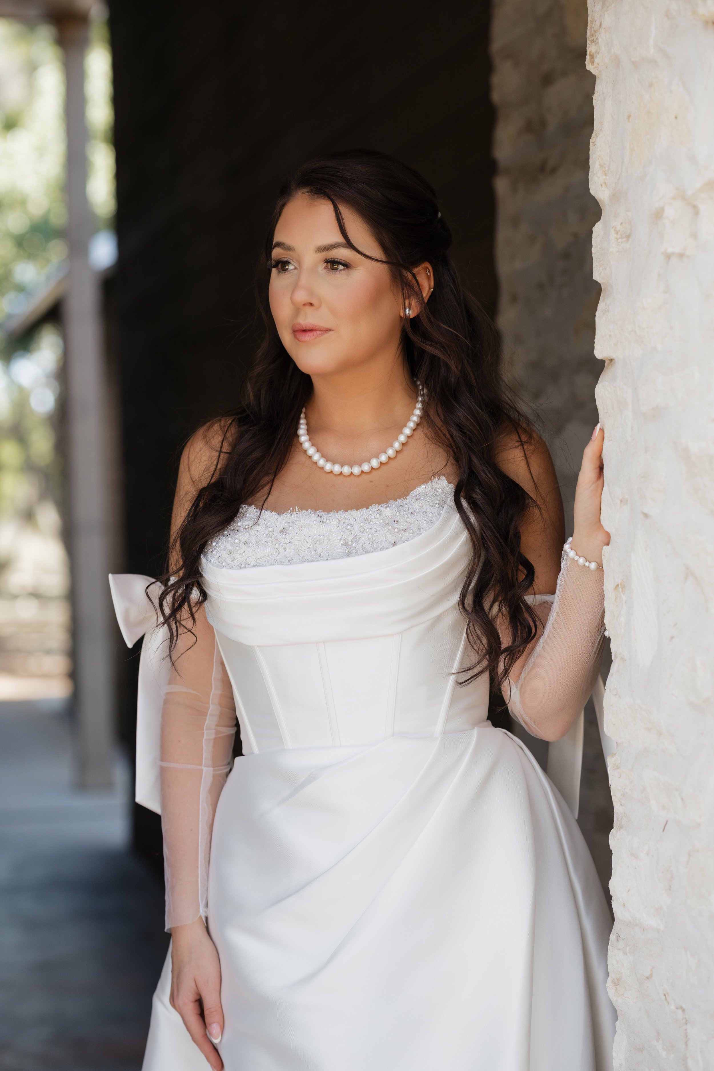 A woman in a wedding dress standing near a stone wall, looking off into the distance.