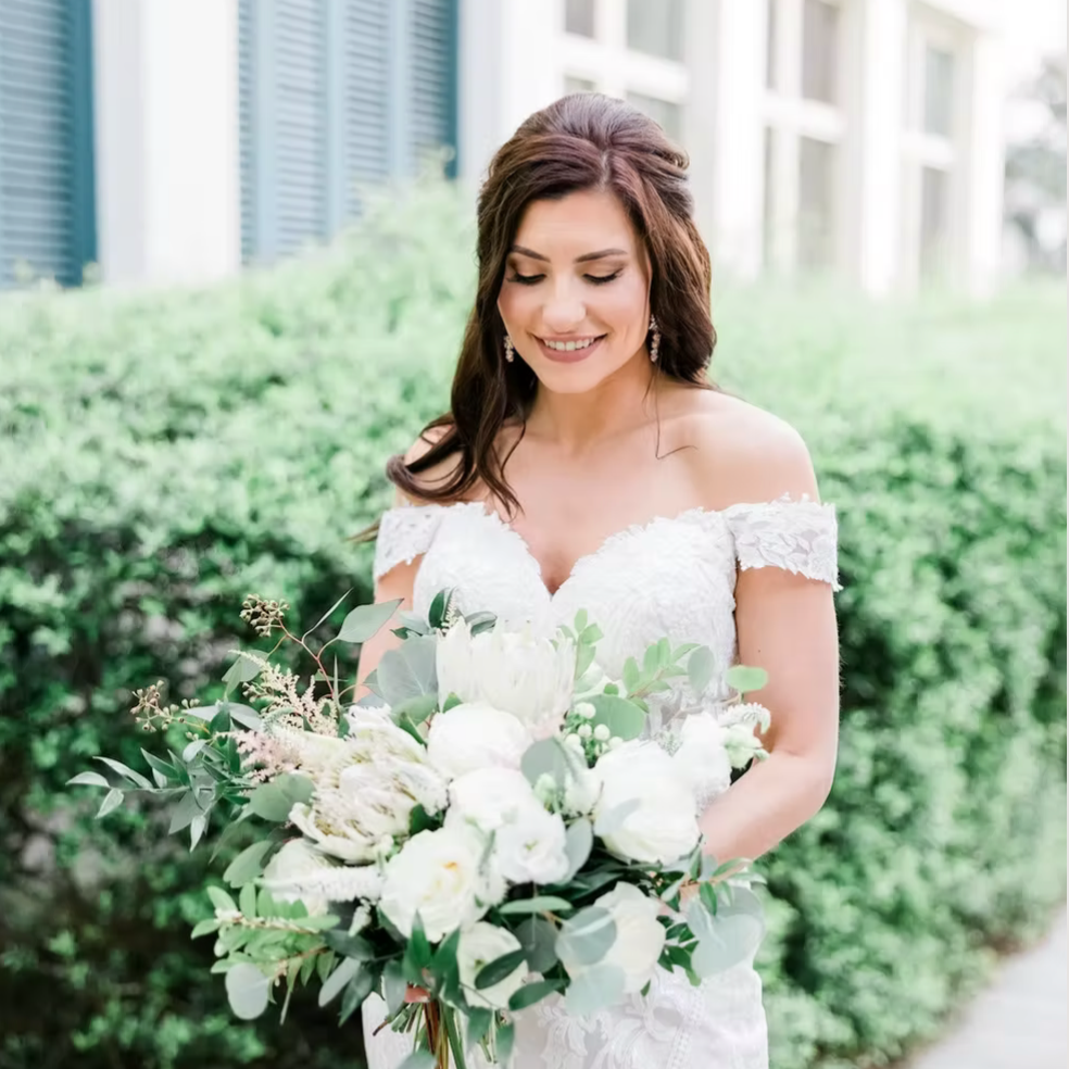 A smiling bride in a white lace wedding dress holding a bouquet of white flowers and greenery outdoors.