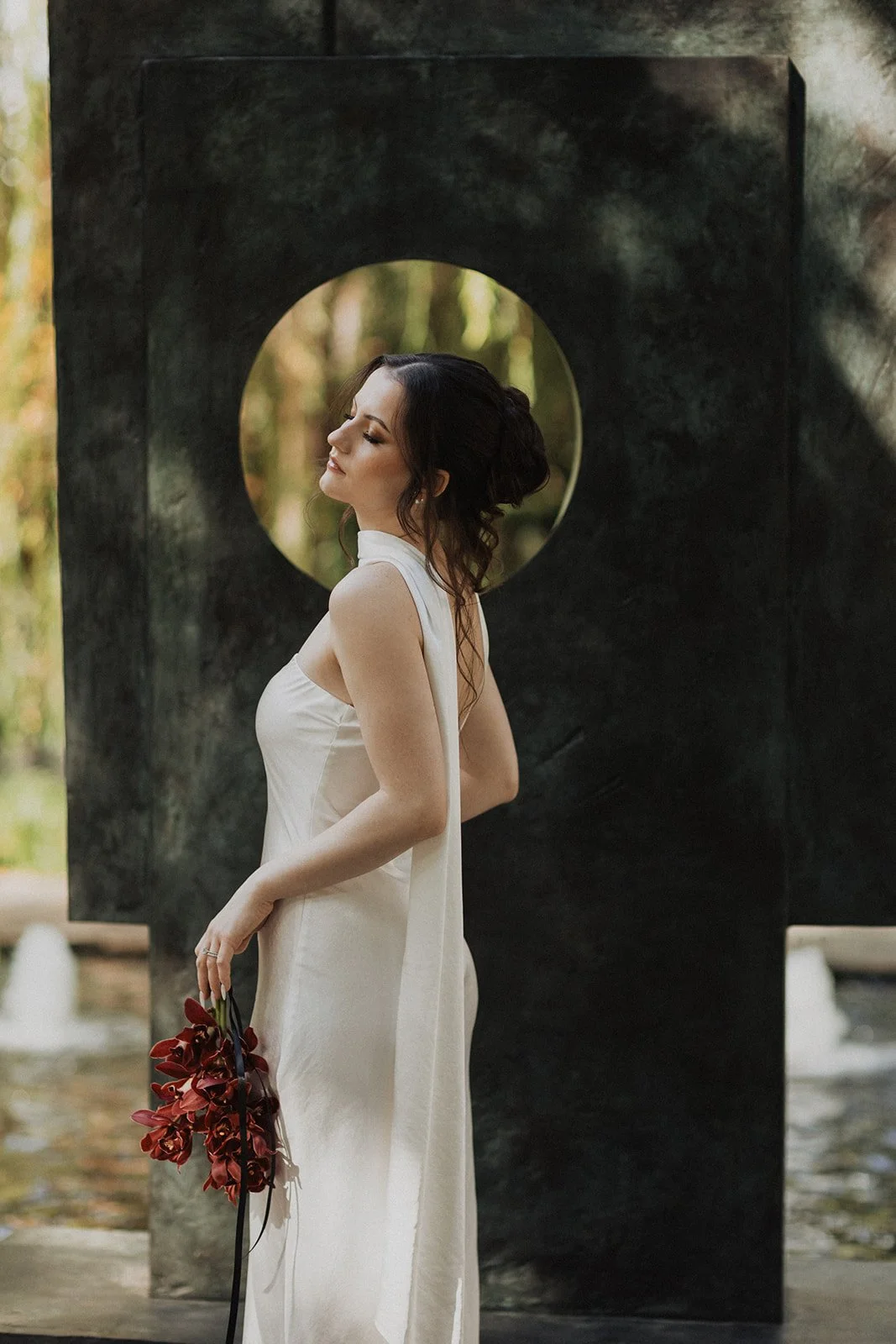 A woman in a white dress holding a bouquet of red flowers standing outdoors beside a black sculpture with a circular cutout.