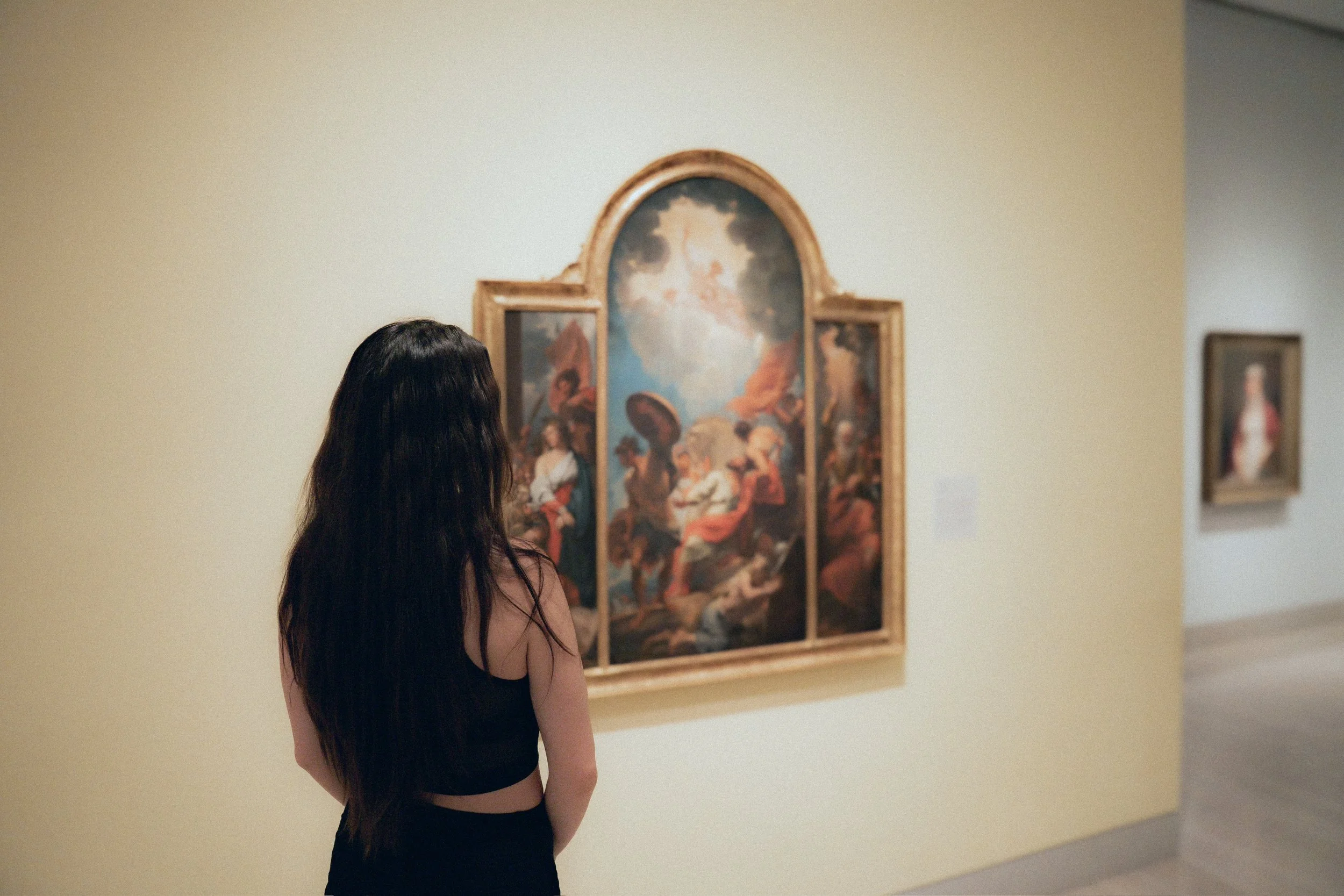 A woman with long dark hair, wearing a black top and pants, observing artwork in an art gallery.