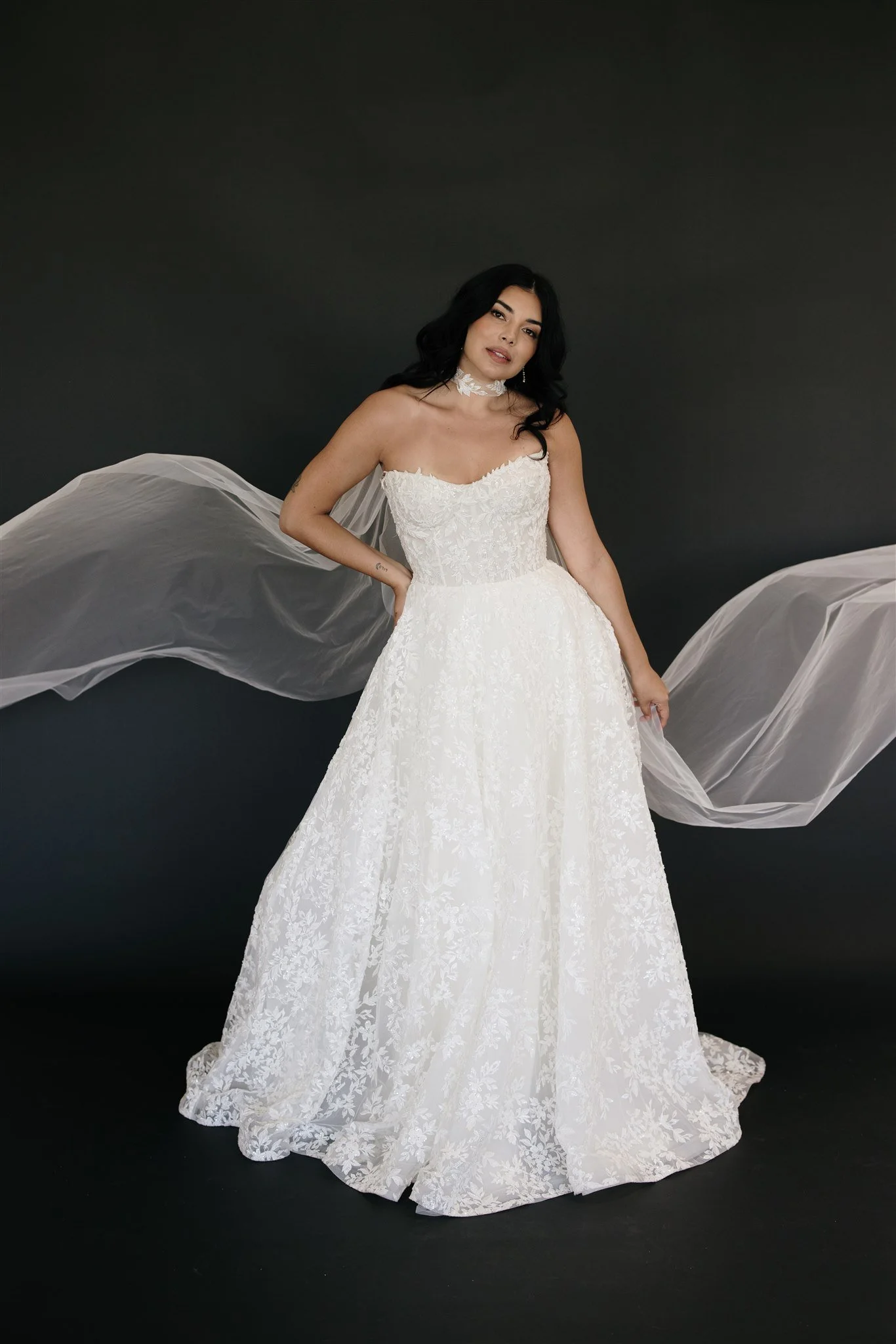 A woman in a white wedding dress with floral lace details, standing against a dark background, with sheer fabric extending from her arms.