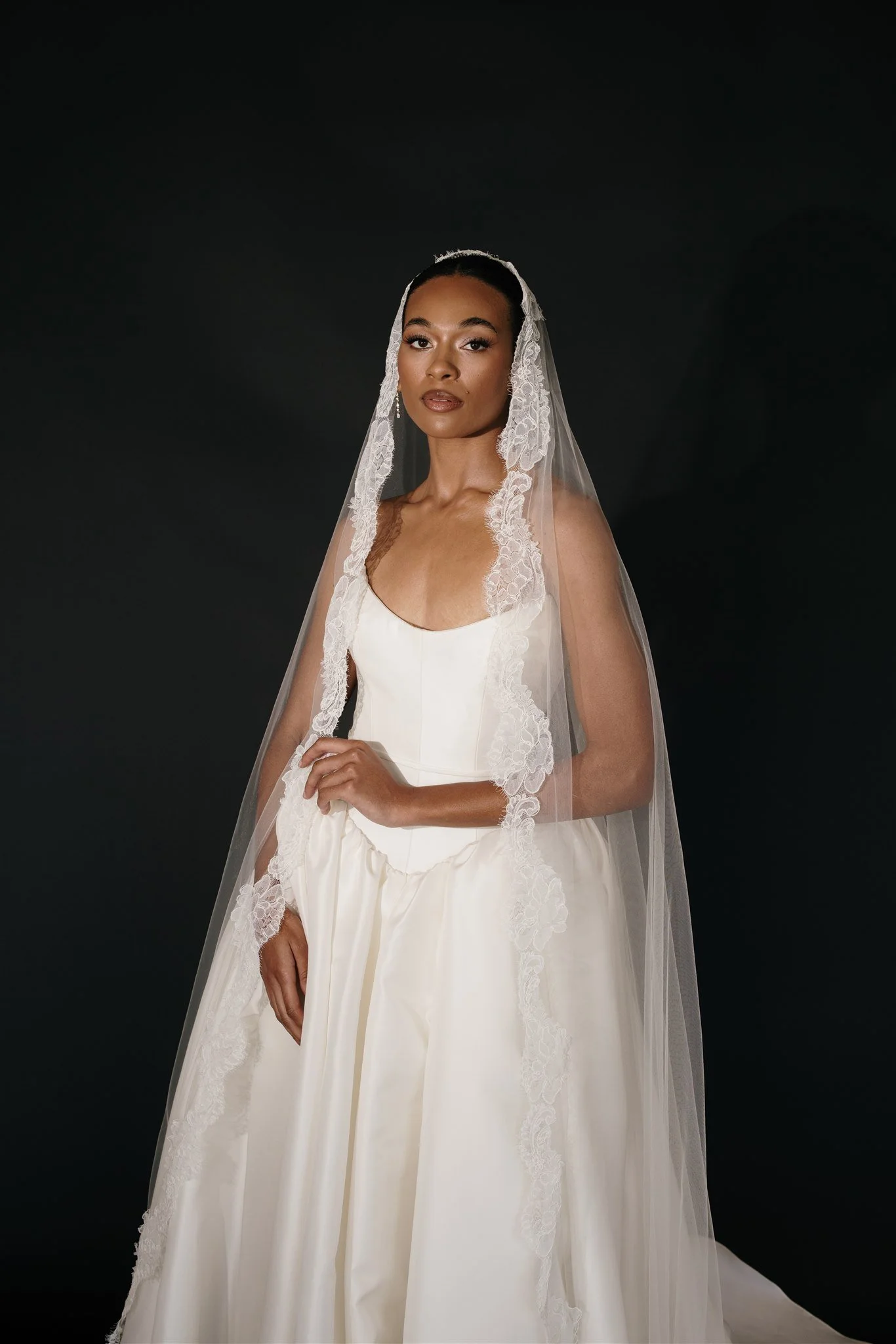 A woman with dark hair in a bun wearing a wedding dress with lace details and a veil, standing against a black background.