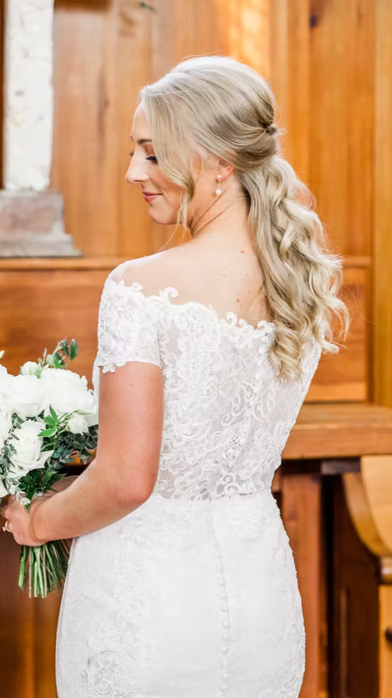 Bride in a white lace wedding dress holding a bouquet of white roses and greenery, standing indoors with a wooden background.