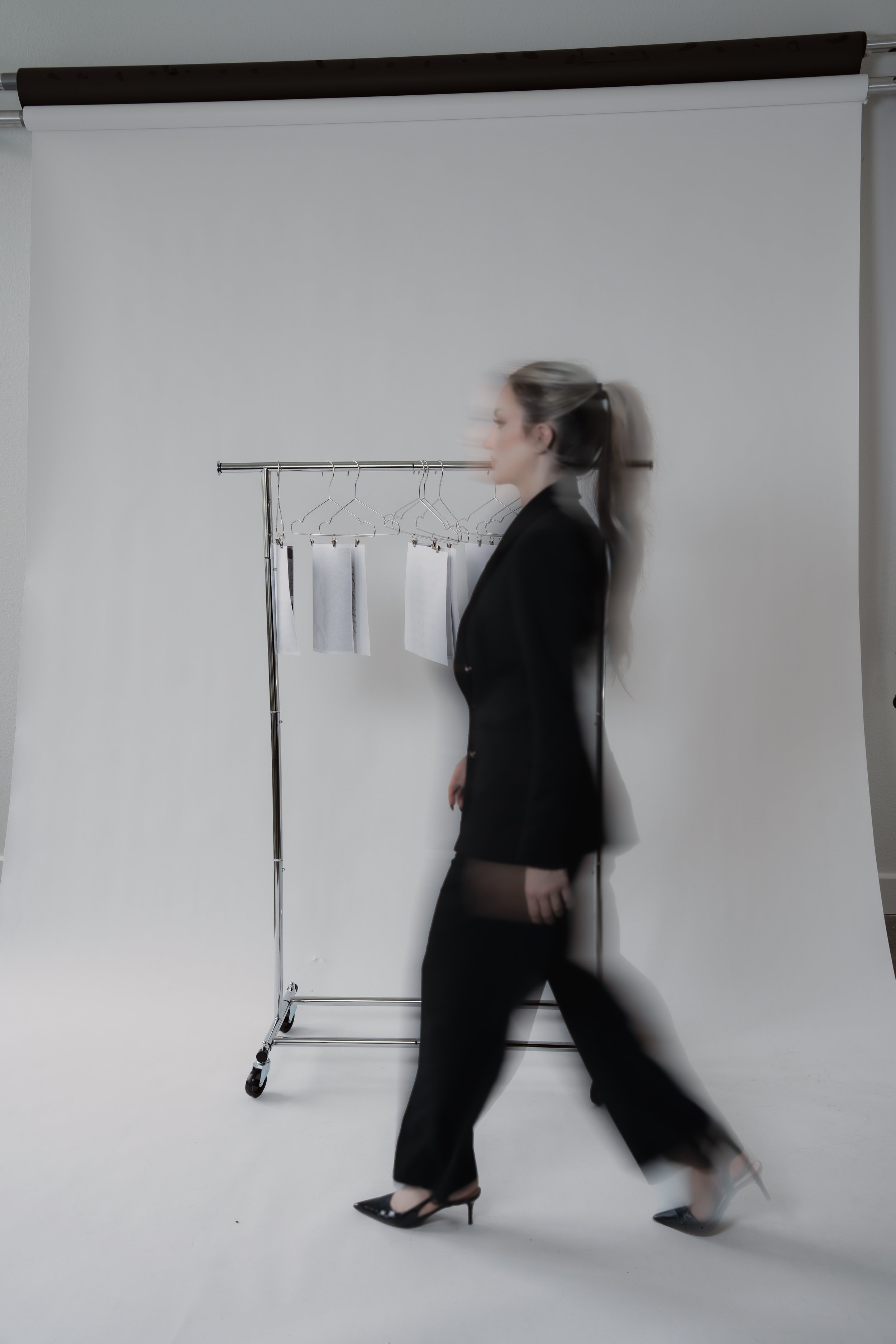 A woman walking past a clothing rack with hangers and white fabric sample swatches, styled in a professional, fashion-focused setting.
