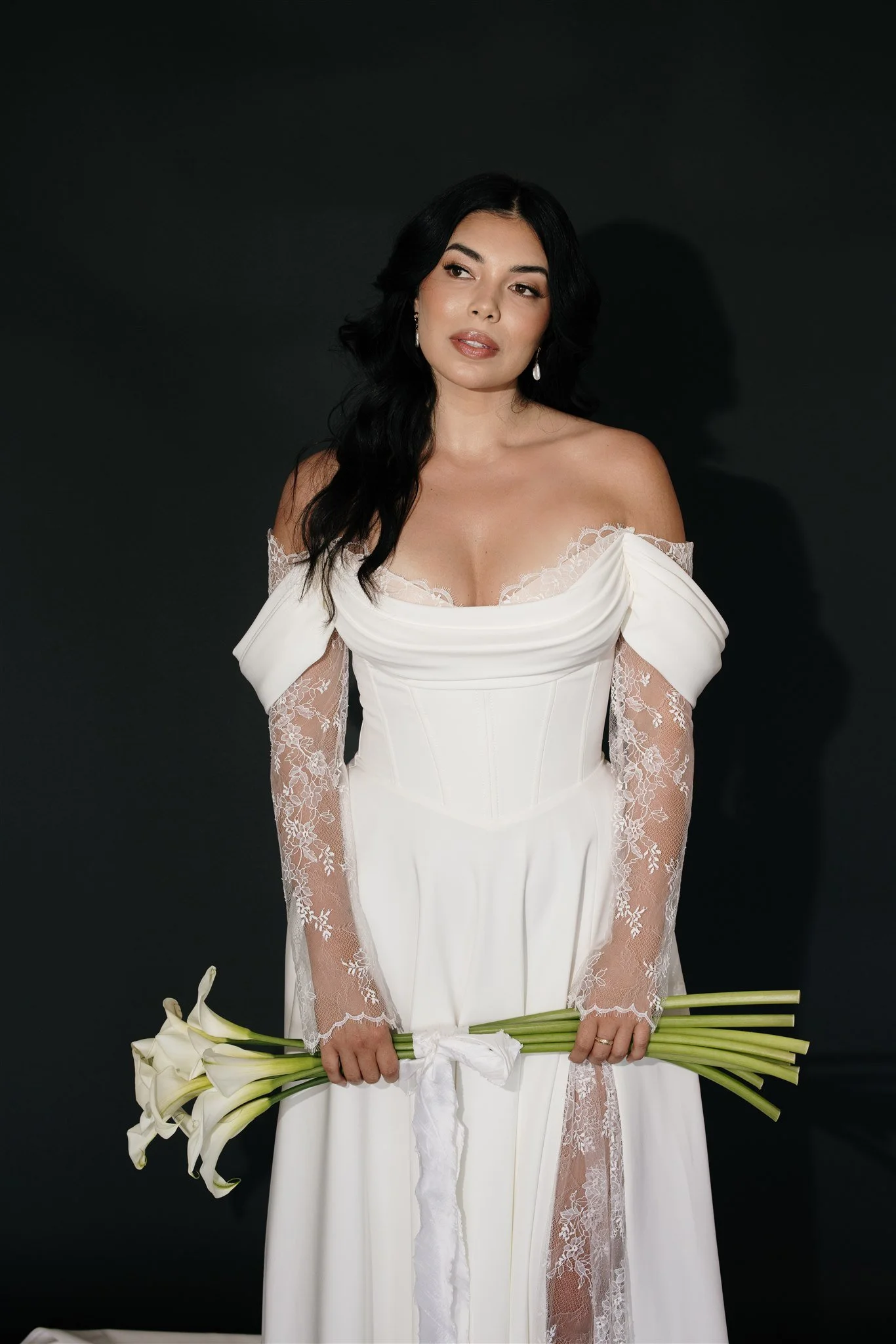 A woman in a white wedding gown with lace sleeves and off-shoulder design holding a bouquet of white calla lilies against a black background.
