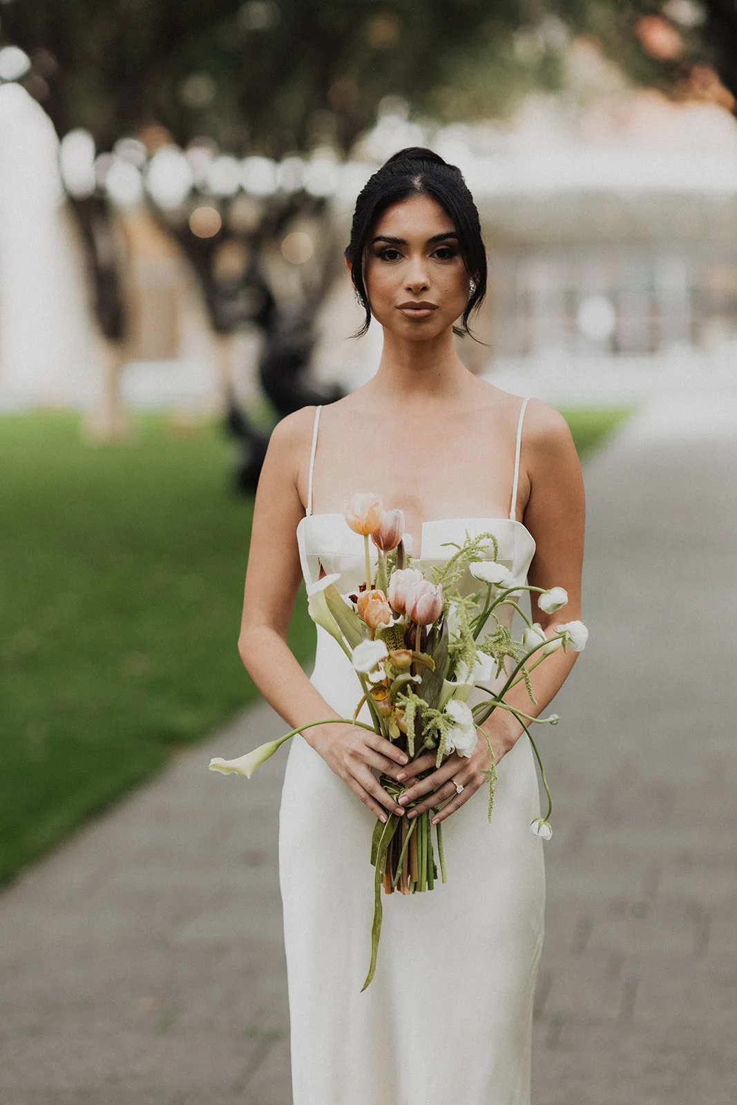 A woman in a white dress holding a bouquet of pink and white flowers outdoors near a body of water with trees and some blurriness in the background.