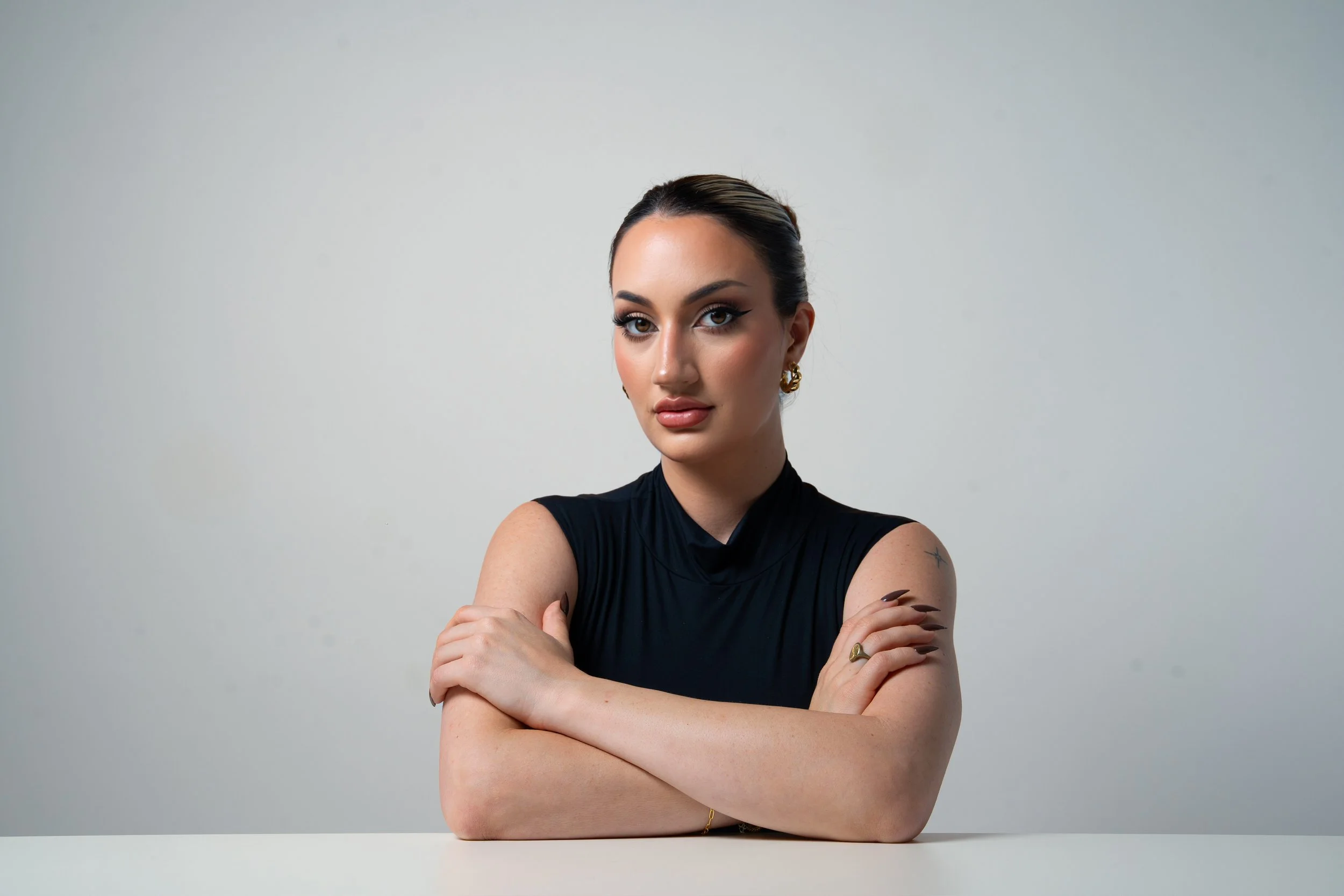 Portrait of a confident woman with crossed arms, wearing a black sleeveless top, gold earrings, and makeup, against a plain light gray background.