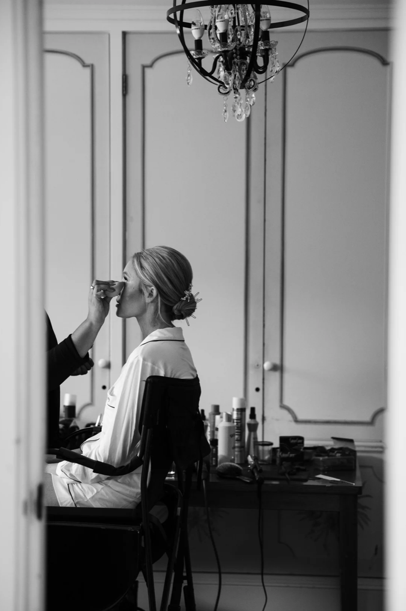 A woman with her hair in an updo sits on a chair as a makeup artist applies makeup to her face in a well-lit room with a chandelier and a large ornate mirror.