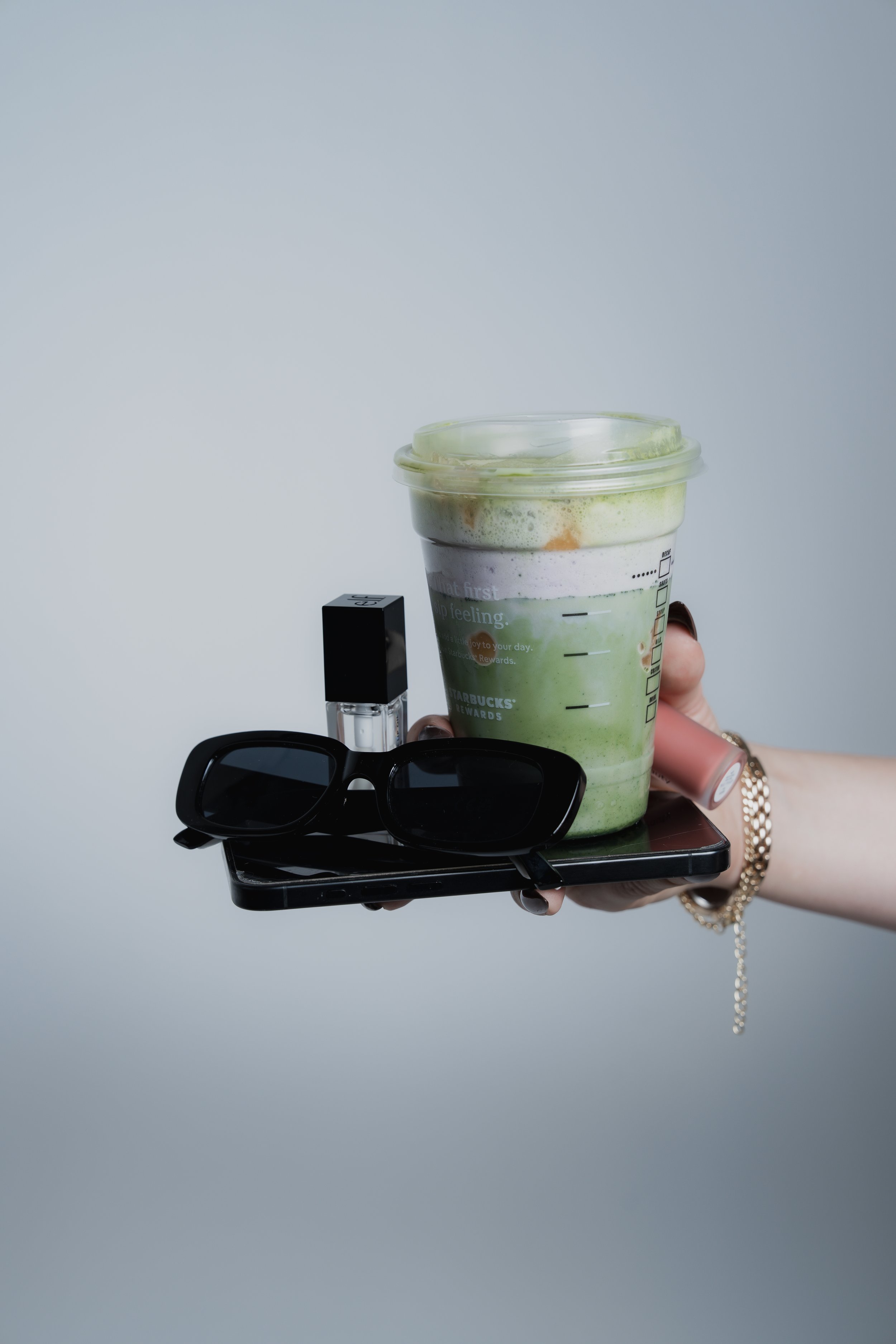 A hand holding a tray with a Starbucks green tea latte and a pair of sunglasses, a lipstick, and a lipstick cap, against a plain white background.
