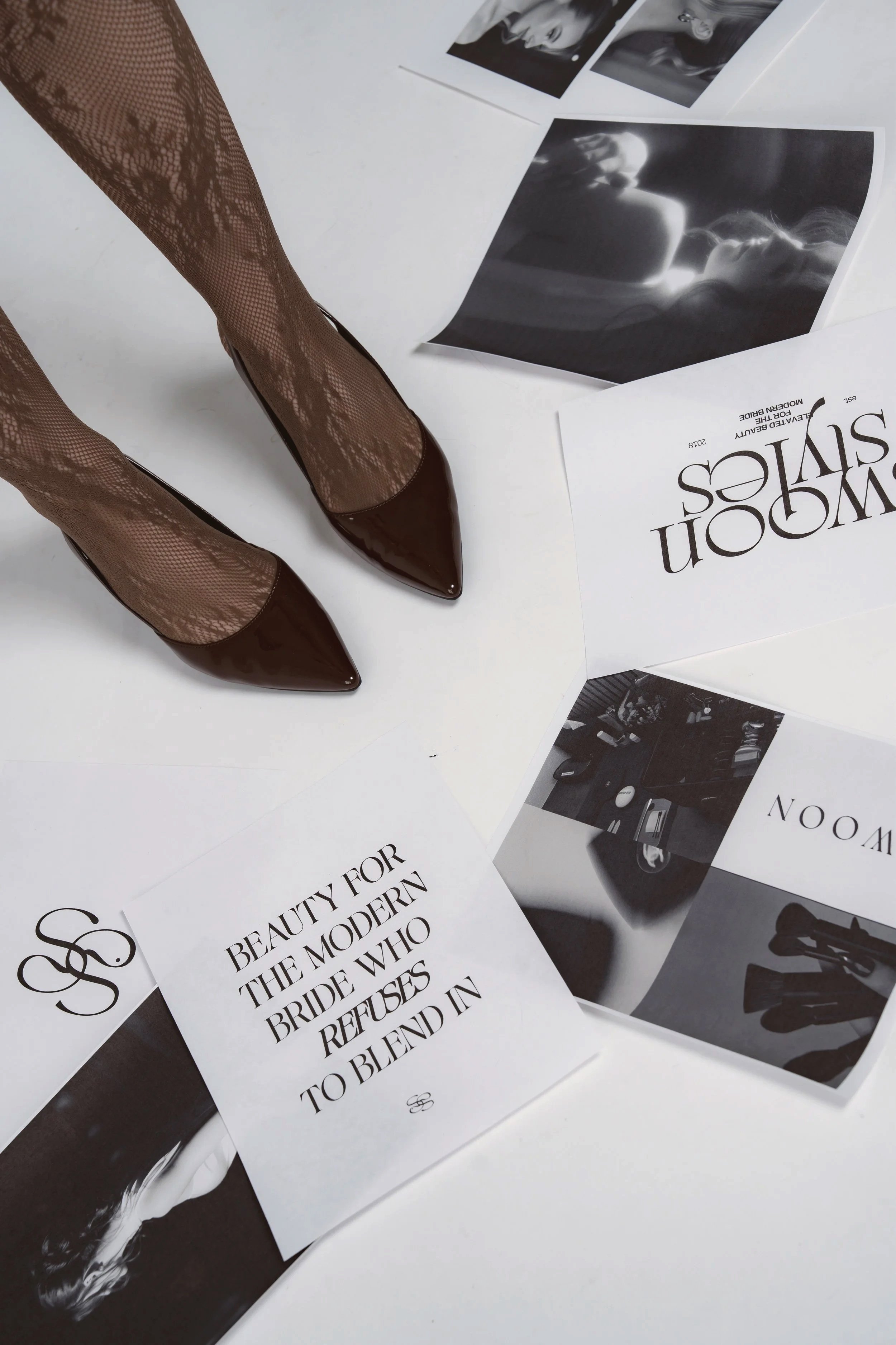Close-up of a pair of legs wearing black fishnet stockings and pointed brown high heels, surrounded by black-and-white magazine pages and photographs on a white surface.