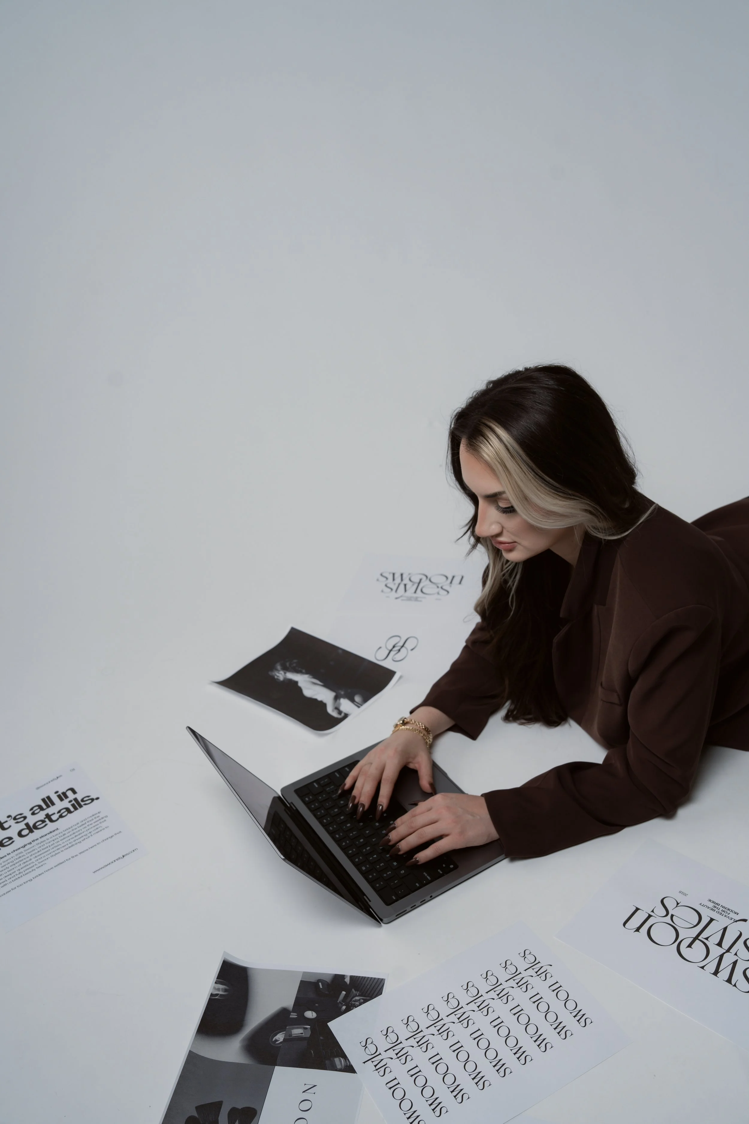 A woman with black and blonde hair sitting at a white table with multiple black and white printouts and a laptop, working or browsing on her laptop.