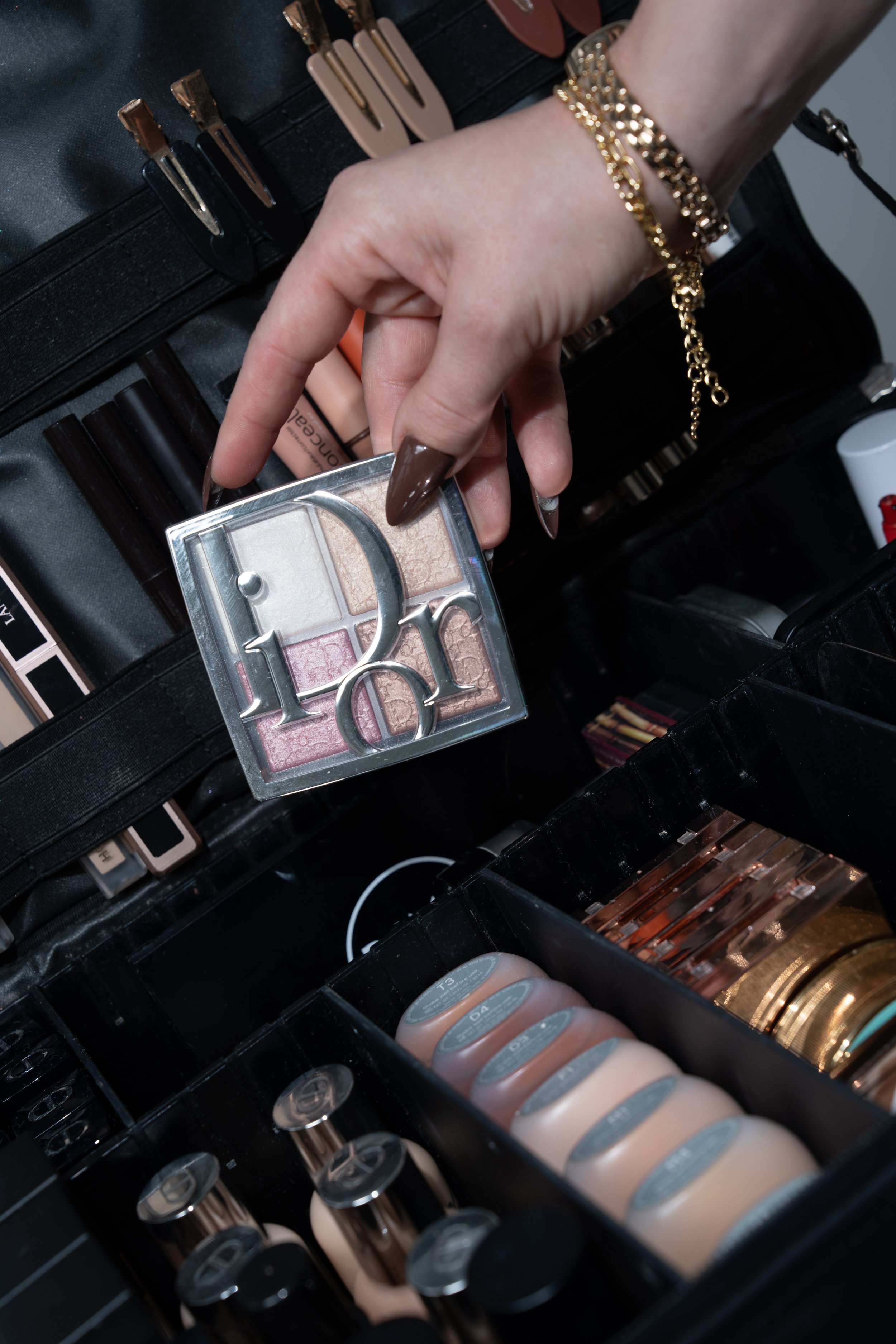 Close-up of a makeup artist's hand with long brown nails, wearing gold jewelry, reaching into a black makeup kit filled with various cosmetic products, including eyeshadows, foundations, and brushes.