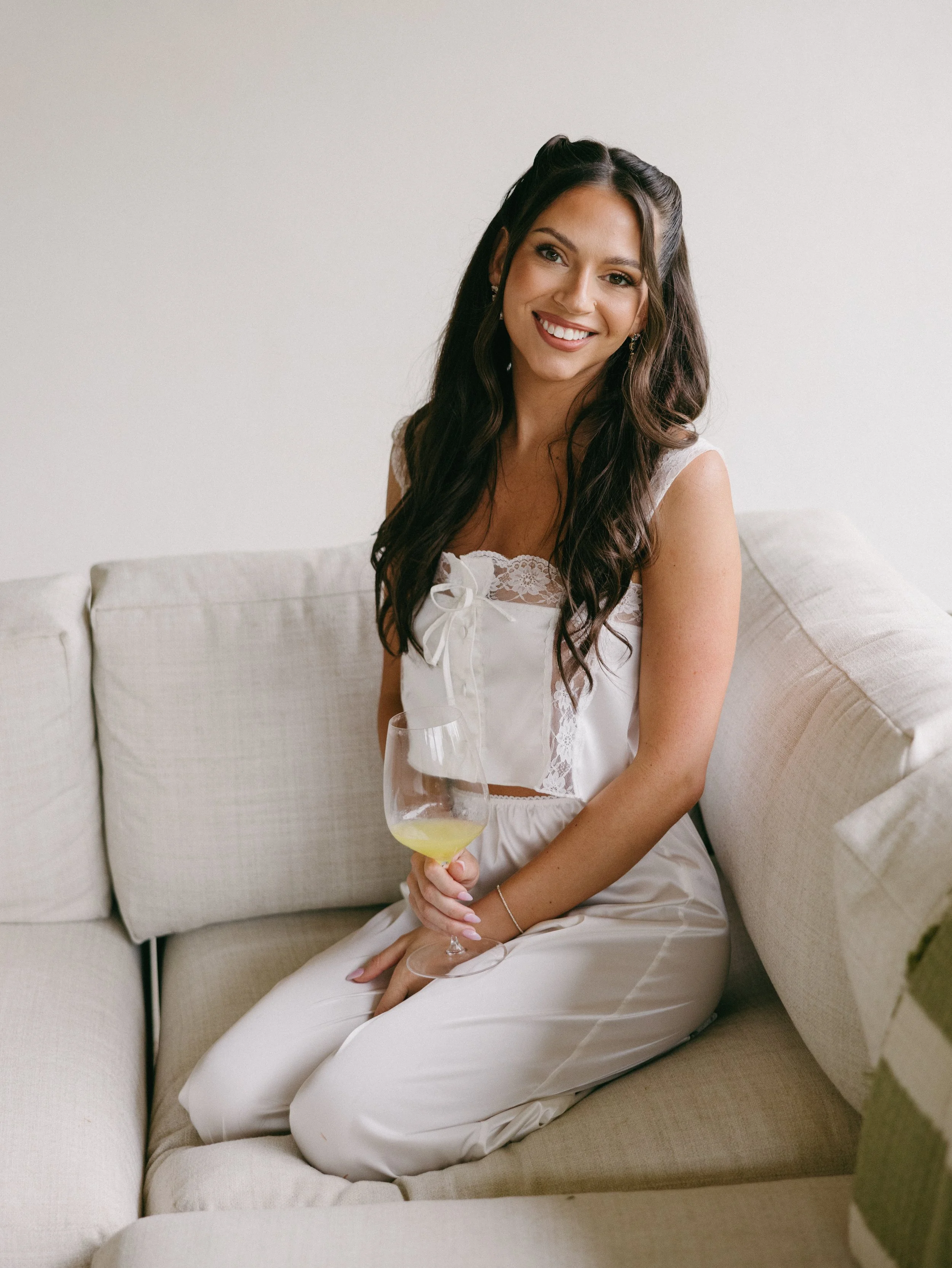 A young woman with long dark wavy hair, smiling, sitting on a beige sofa in a white lace top and matching pants, holding a glass of white wine, in a bright room with a plain white wall.