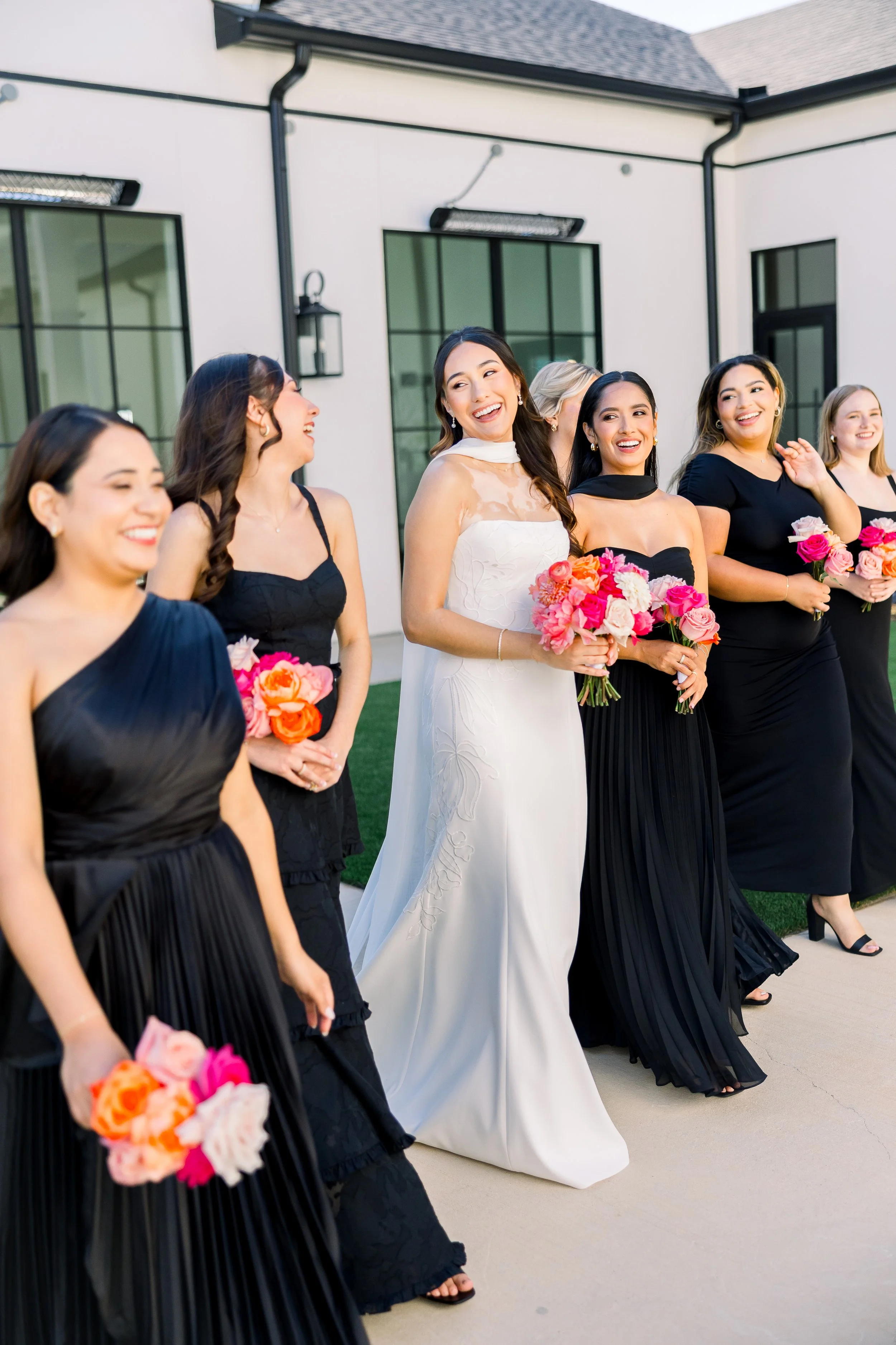 A bride dressed in a white gown with sheer details on the bodice standing with bridesmaids dressed in black, holding bouquets of pink and coral flowers, as they walk outdoors near a modern building with large windows and black trim.