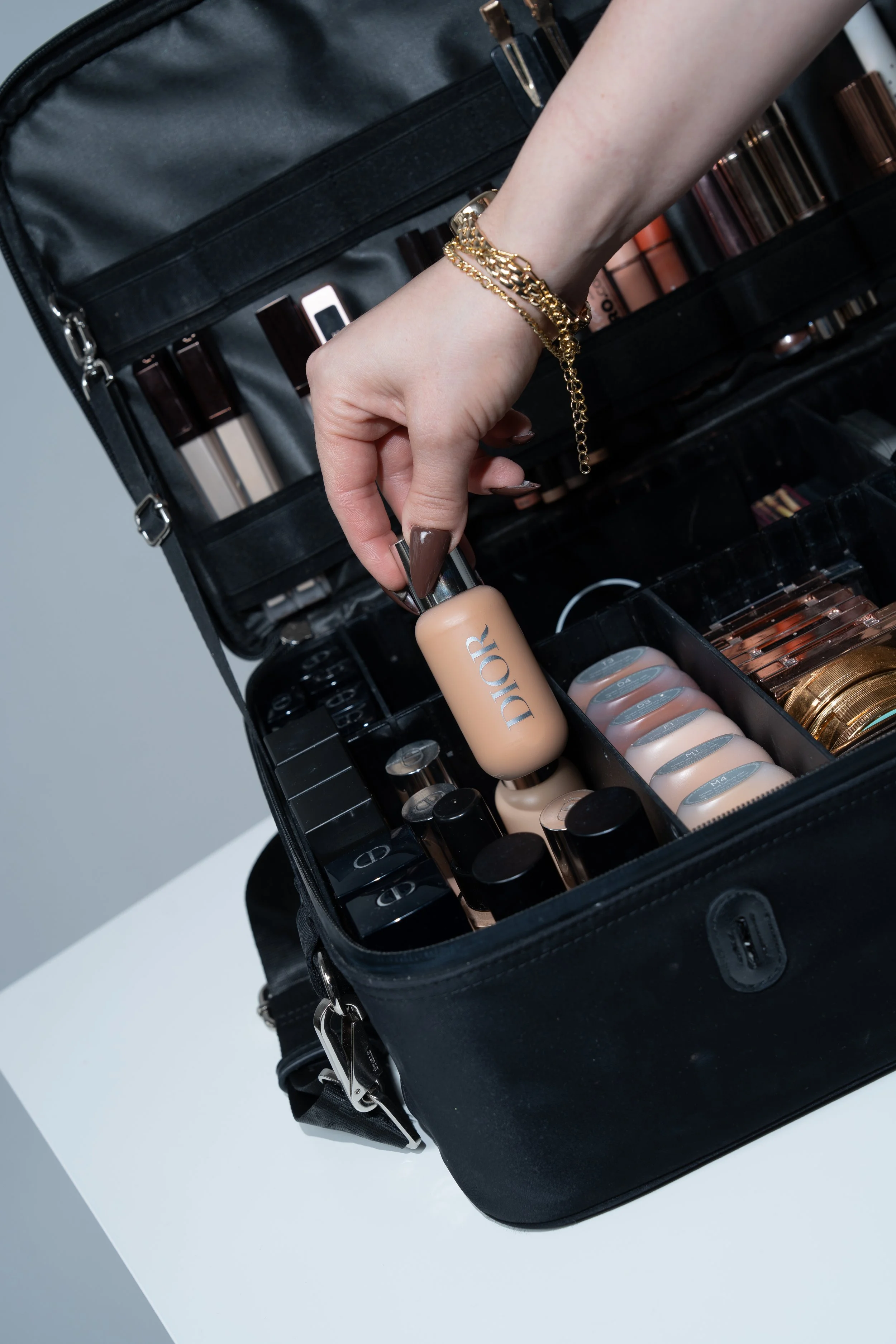 A hand with gold jewelry reaching into a black makeup case, which contains foundation bottles, lip glosses, and other makeup products.