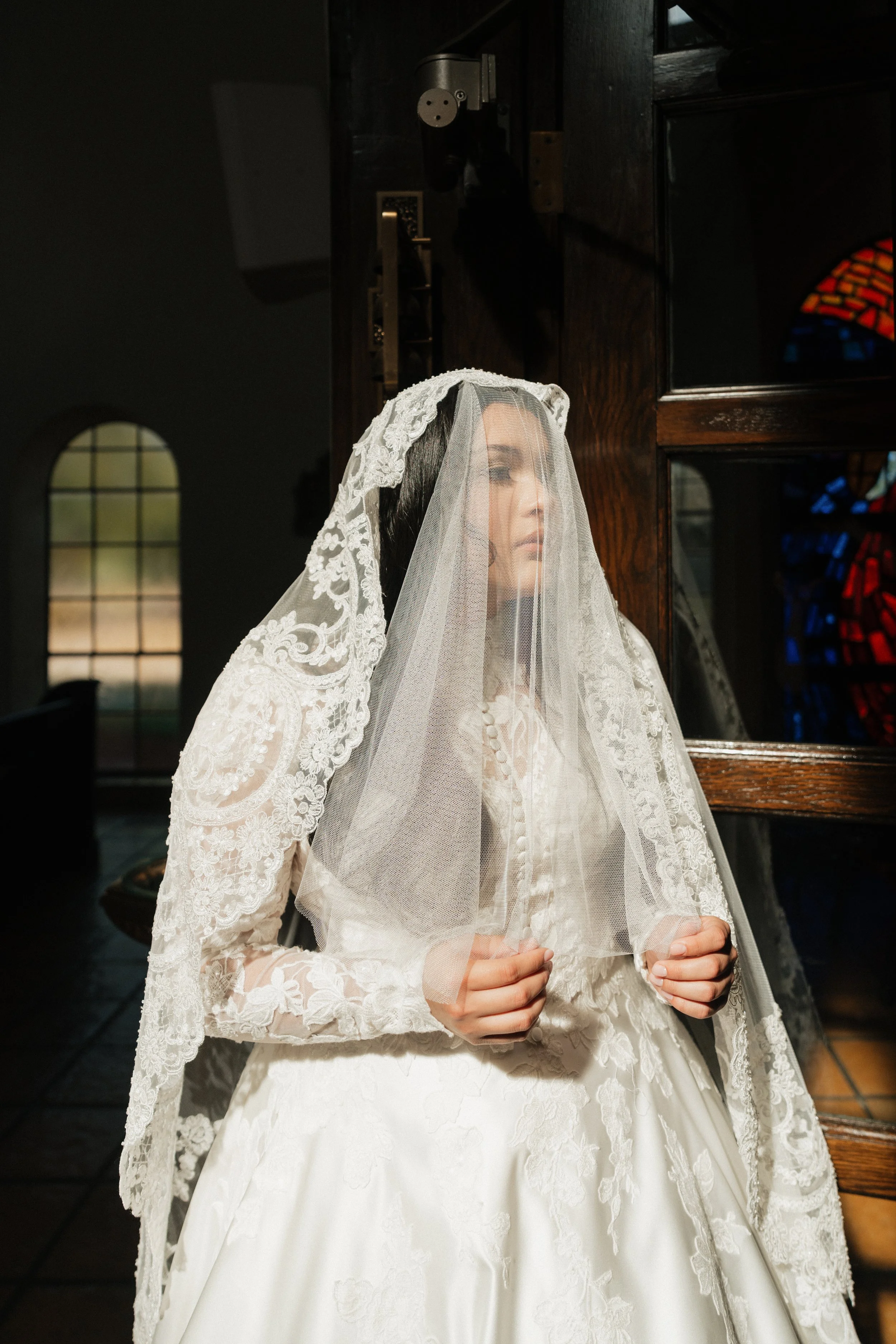 A woman dressed in a white wedding gown with lace details and a lace veil, standing indoors near a wooden cabinet and a window with stained glass, with her hands clasped.
