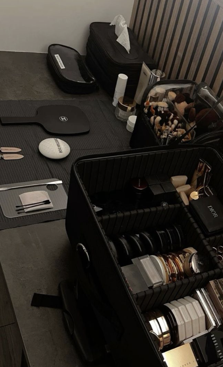 A table filled with makeup products, brushes, and accessories, including a Dior powder compact, lip glosses, and a makeup organizer with various cosmetics. There are also bags and containers in the background.