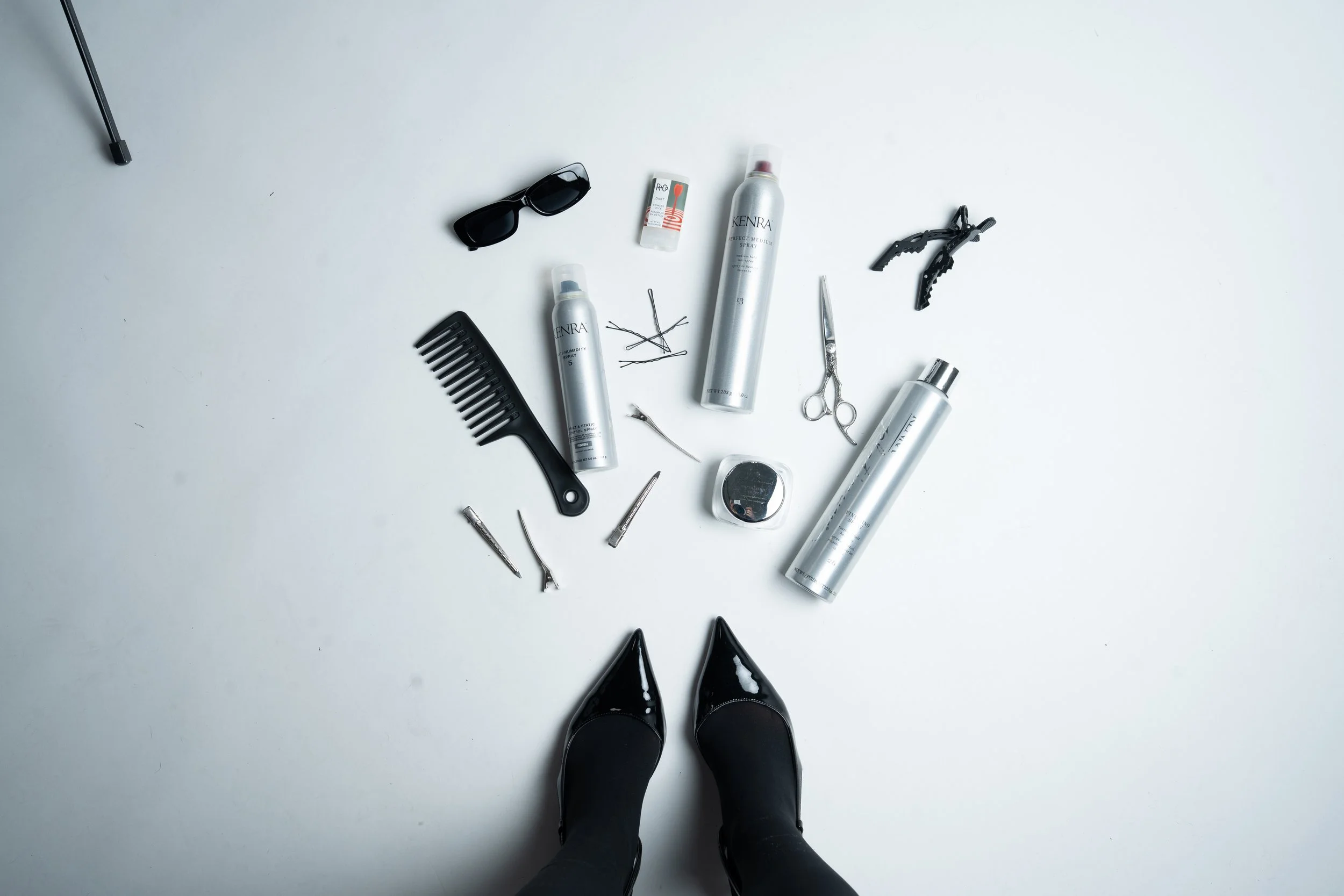 Flat lay of a person in black high heels standing on a white surface surrounded by hairstyling and beauty tools, including hairspray bottles, a comb, hairpins, scissors, clamps, and sunglasses.