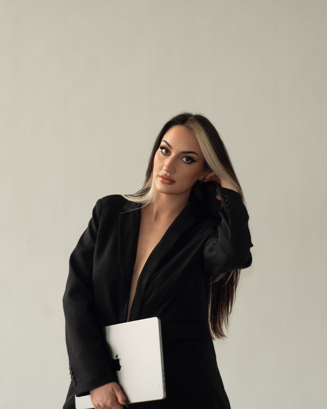 A woman with long dark hair and blonde streaks, wearing a black blazer, holding a silver laptop, standing against a plain light background.