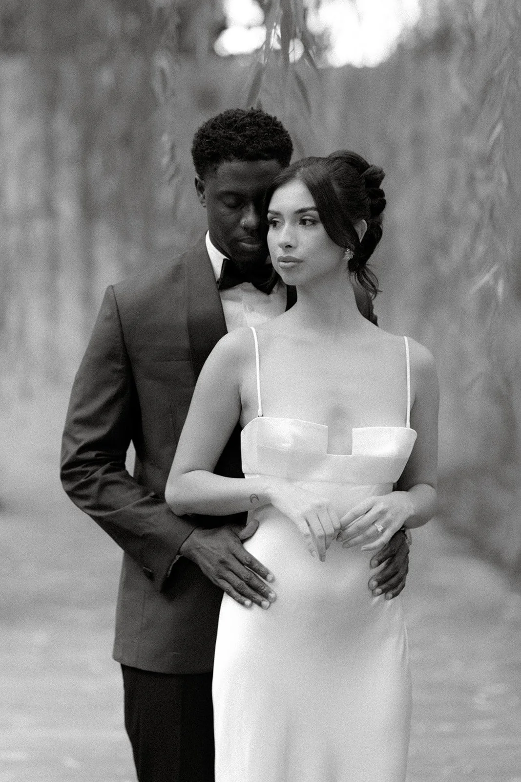 A black-and-white photo of a man and woman in formal attire, standing close together outdoors. The man is wearing a tuxedo and the woman is in a white dress with thin straps.
