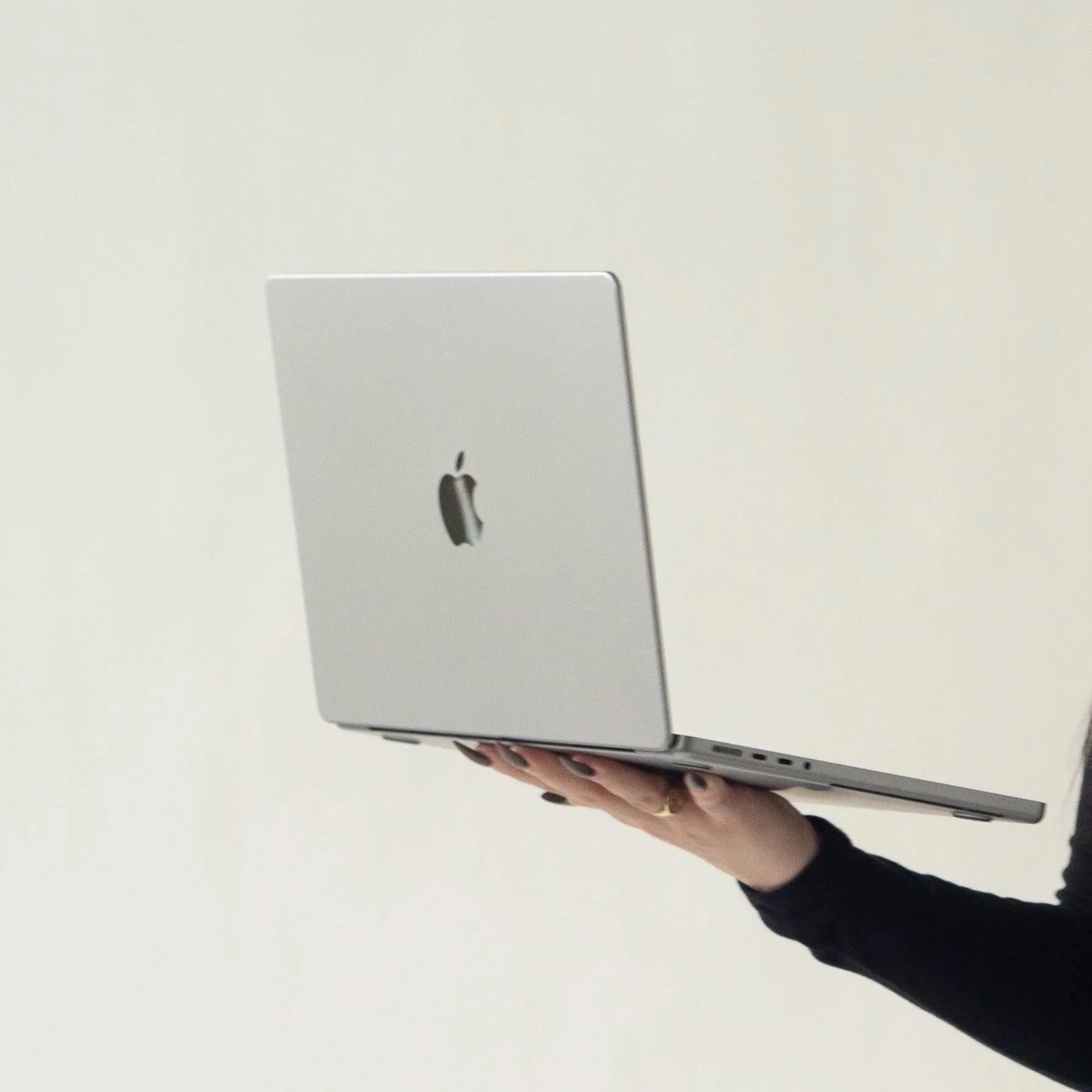 A person holding a silver MacBook laptop with an Apple logo, against a plain light-colored background.