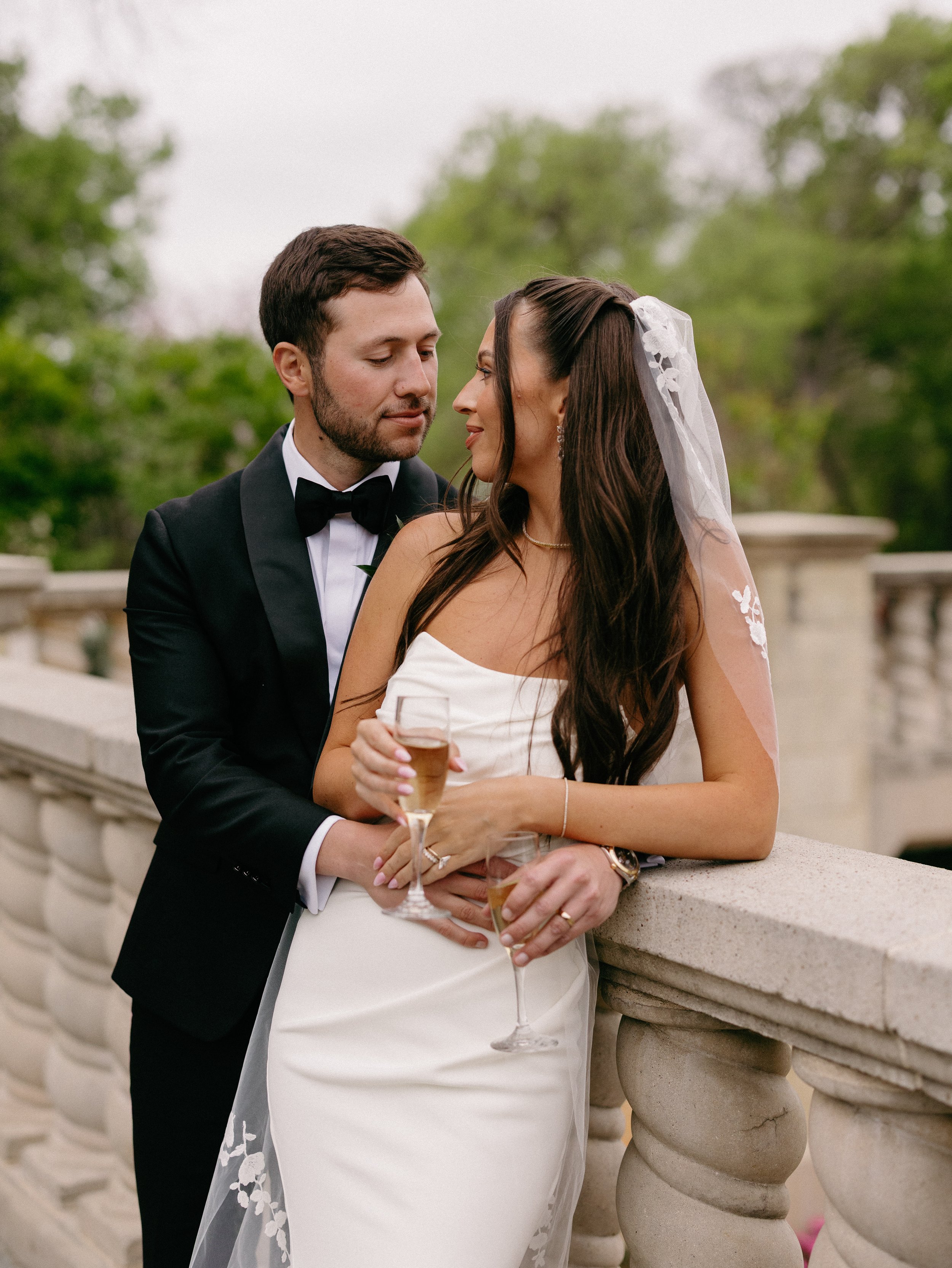 A bride and groom on a balcony, with the groom standing behind the bride. The groom is wearing a black tuxedo with a bow tie, and the bride is in a strapless white wedding dress holding a champagne glass. They are looking at each other affectionately, with a blurred background of green trees.