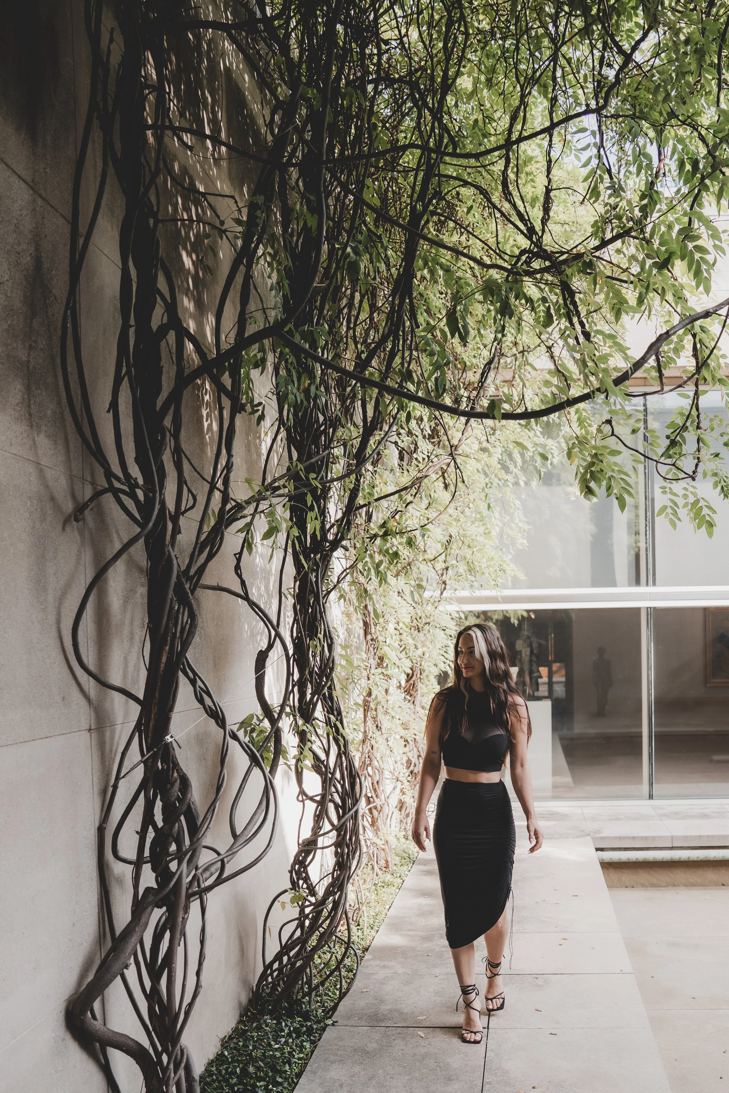 A woman with long dark hair wearing a black sleeveless top and a black skirt with a slit, walking on a concrete sidewalk next to a wall covered in twisting vines. The background features large windows with reflections of an outdoor scene and some trees.