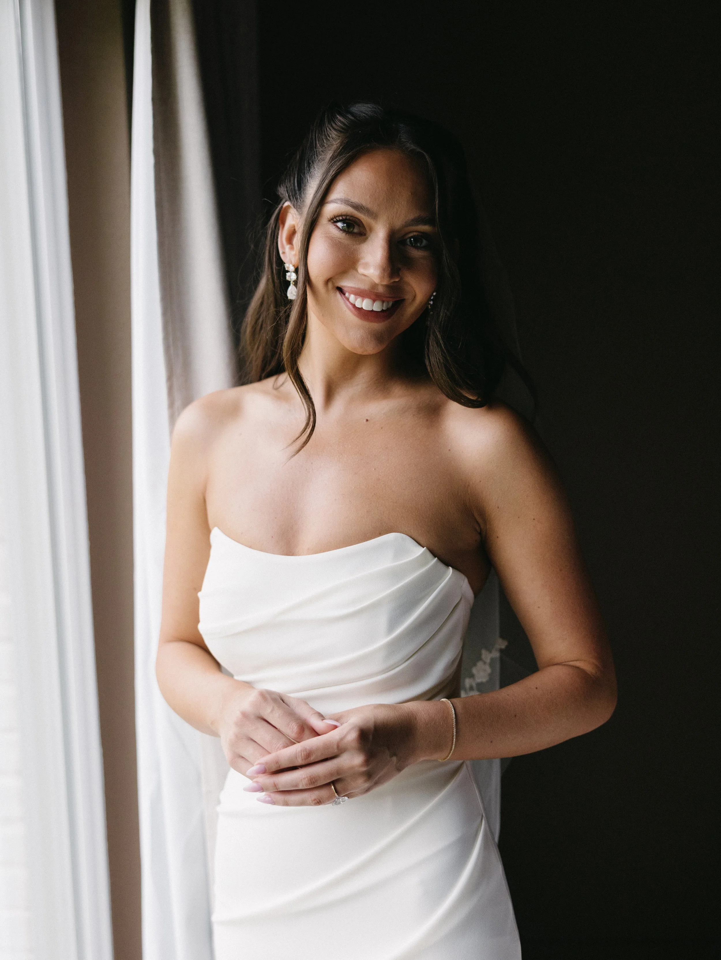 A smiling woman with long dark hair and earrings, wearing a strapless white dress, stands near a window with curtains.