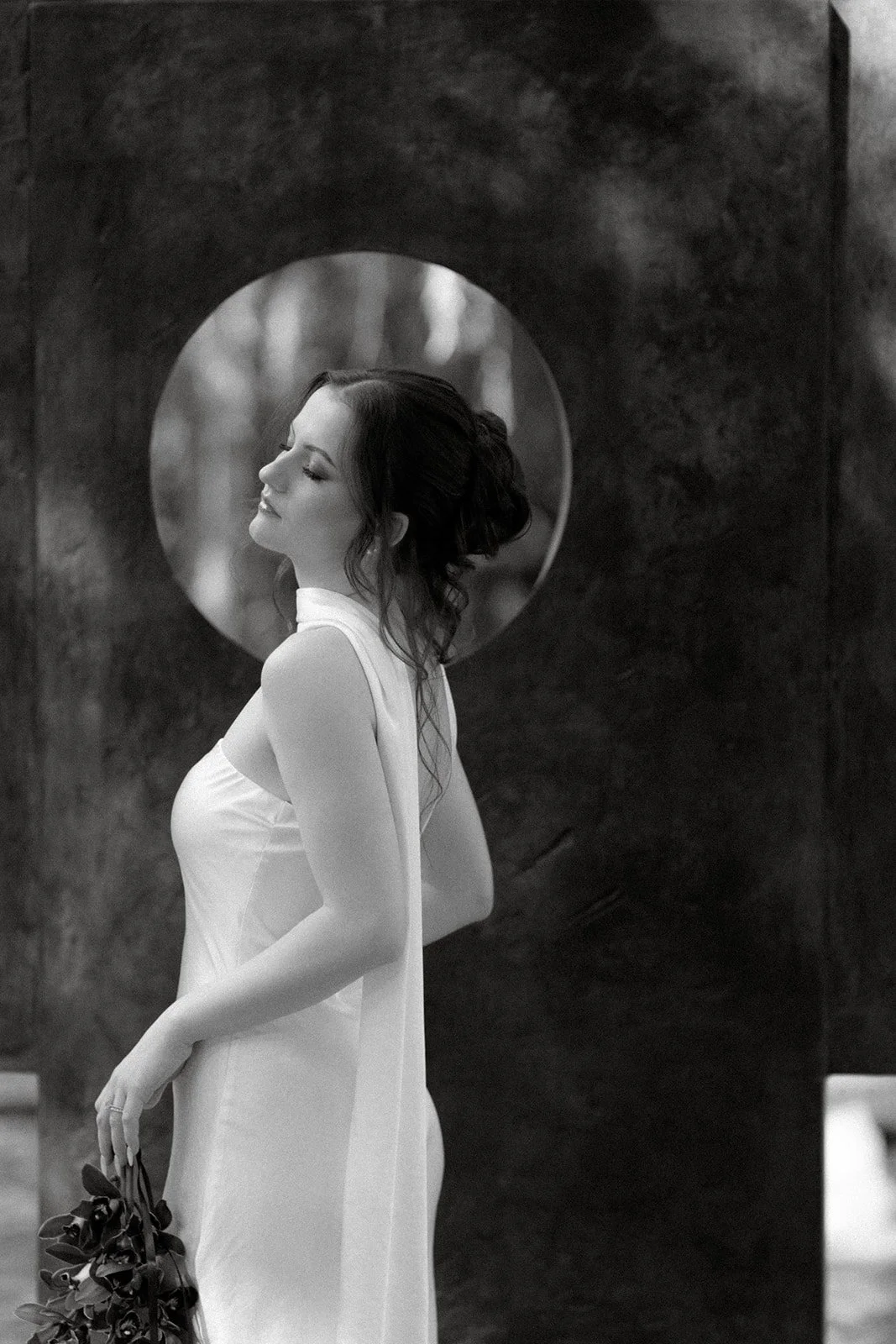 A woman with dark hair styled in loose curls, wearing a sleeveless white dress, stands in profile with her eyes closed, holding a bunch of dark flowers, and smiling softly in front of a round window in a dark wall.