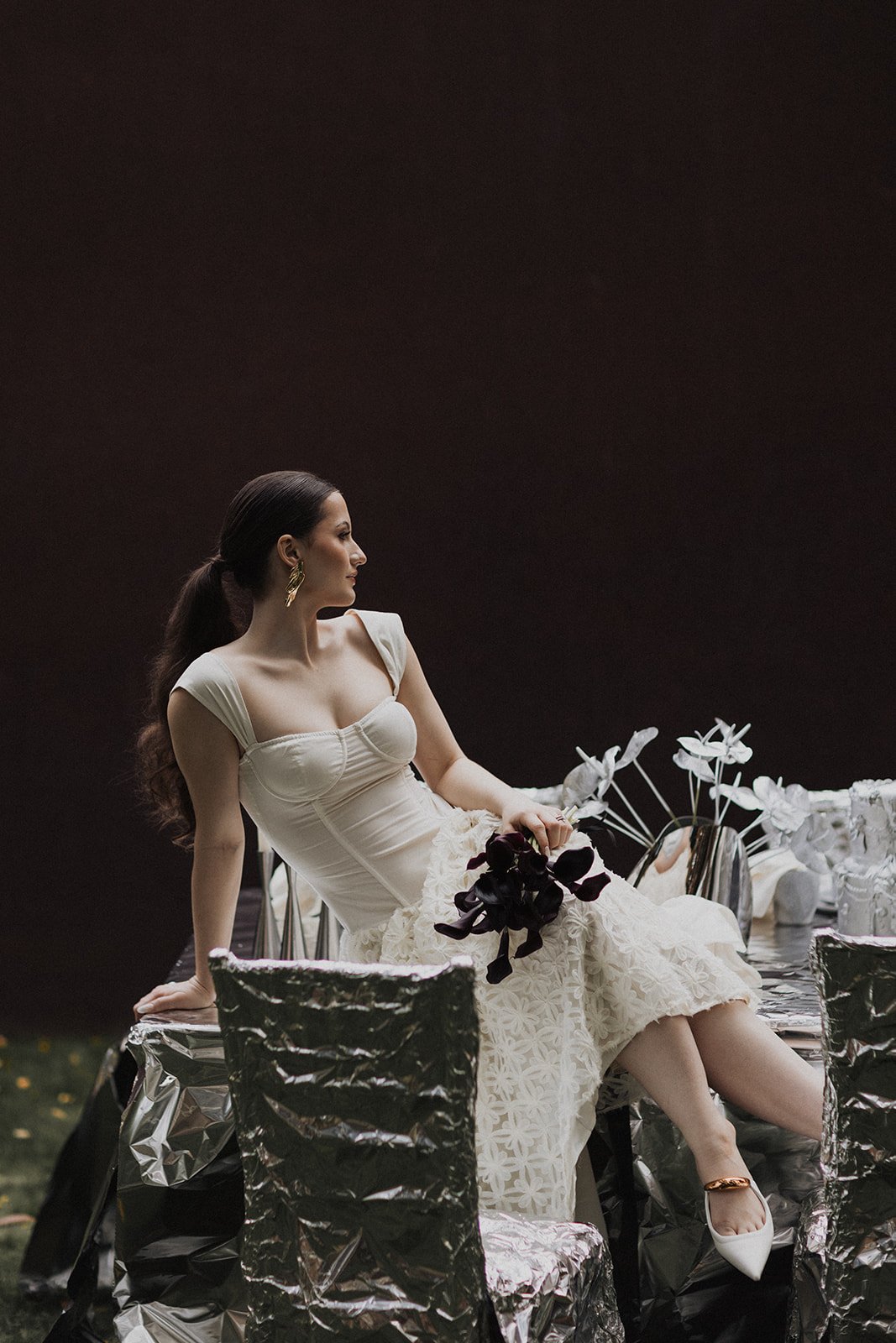 A woman in a white dress sitting on a silver table with black and white flowers and black chairs, against a dark background.