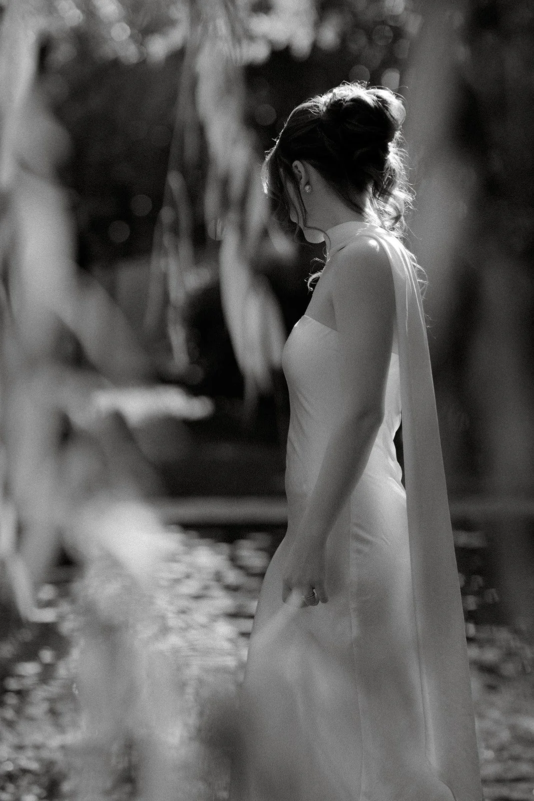 A black and white photo of a woman with styled hair, wearing a strapless dress with a sash, standing outdoors near a body of water, surrounded by natural scenery.