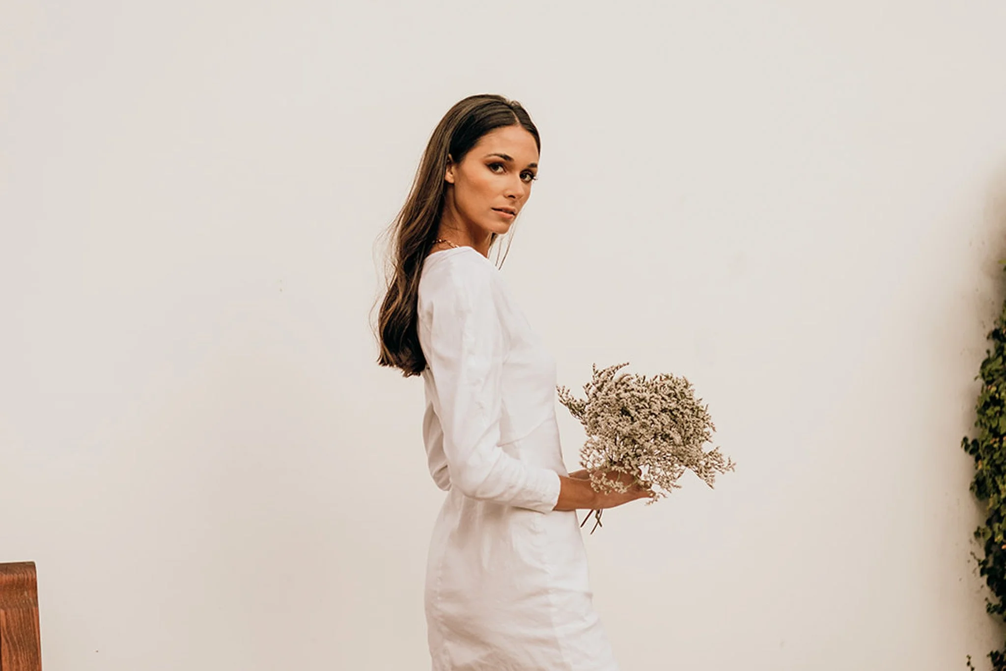 A woman with long brown hair wearing a white outfit holding a bouquet of dried flowers, posed against a plain light-colored wall with some plants on the side.