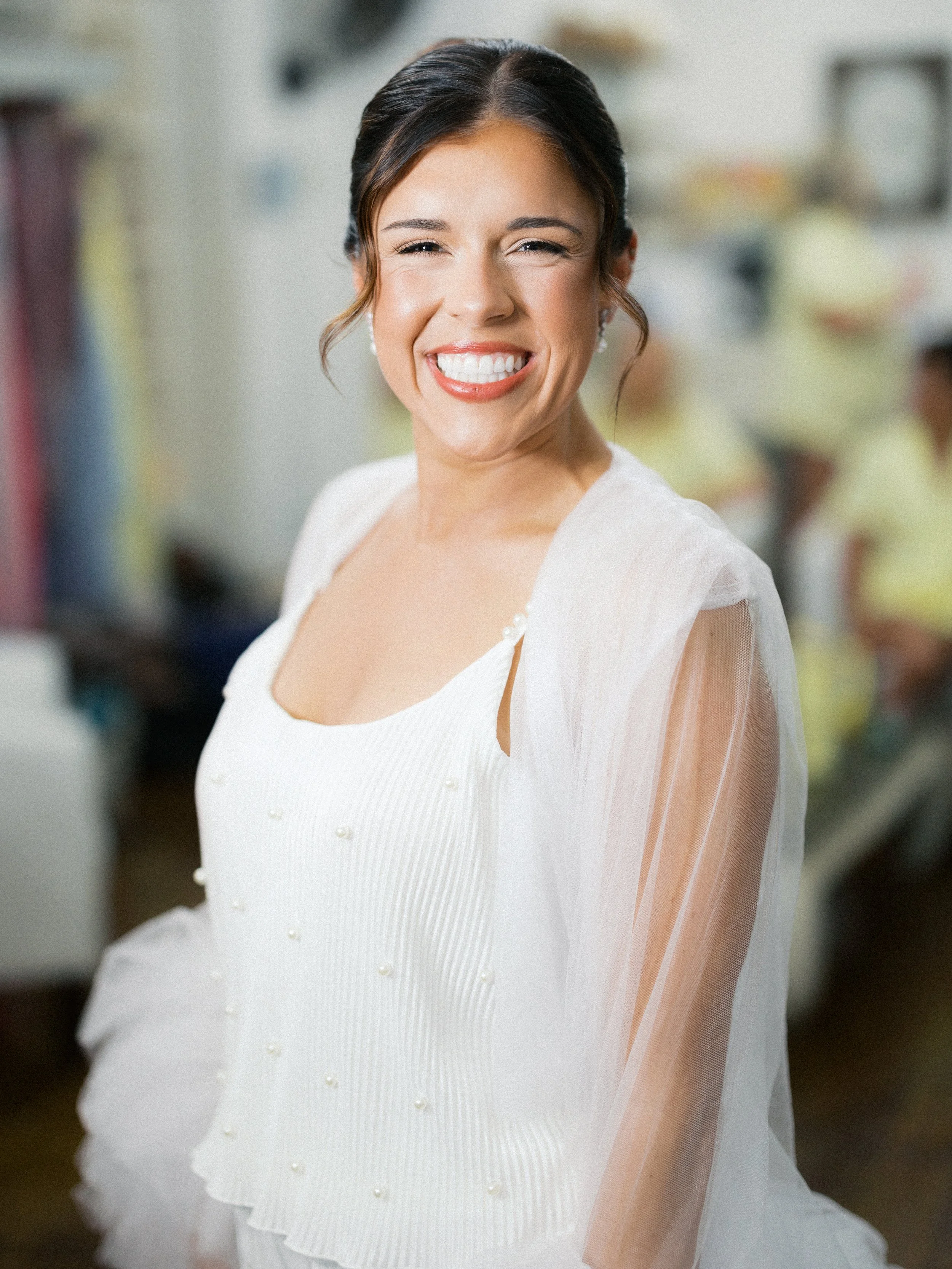A woman smiling while looking at the camera, wearing a white top with pearl embellishments and a sheer cape, standing in a room with blurred background of people and shelves.