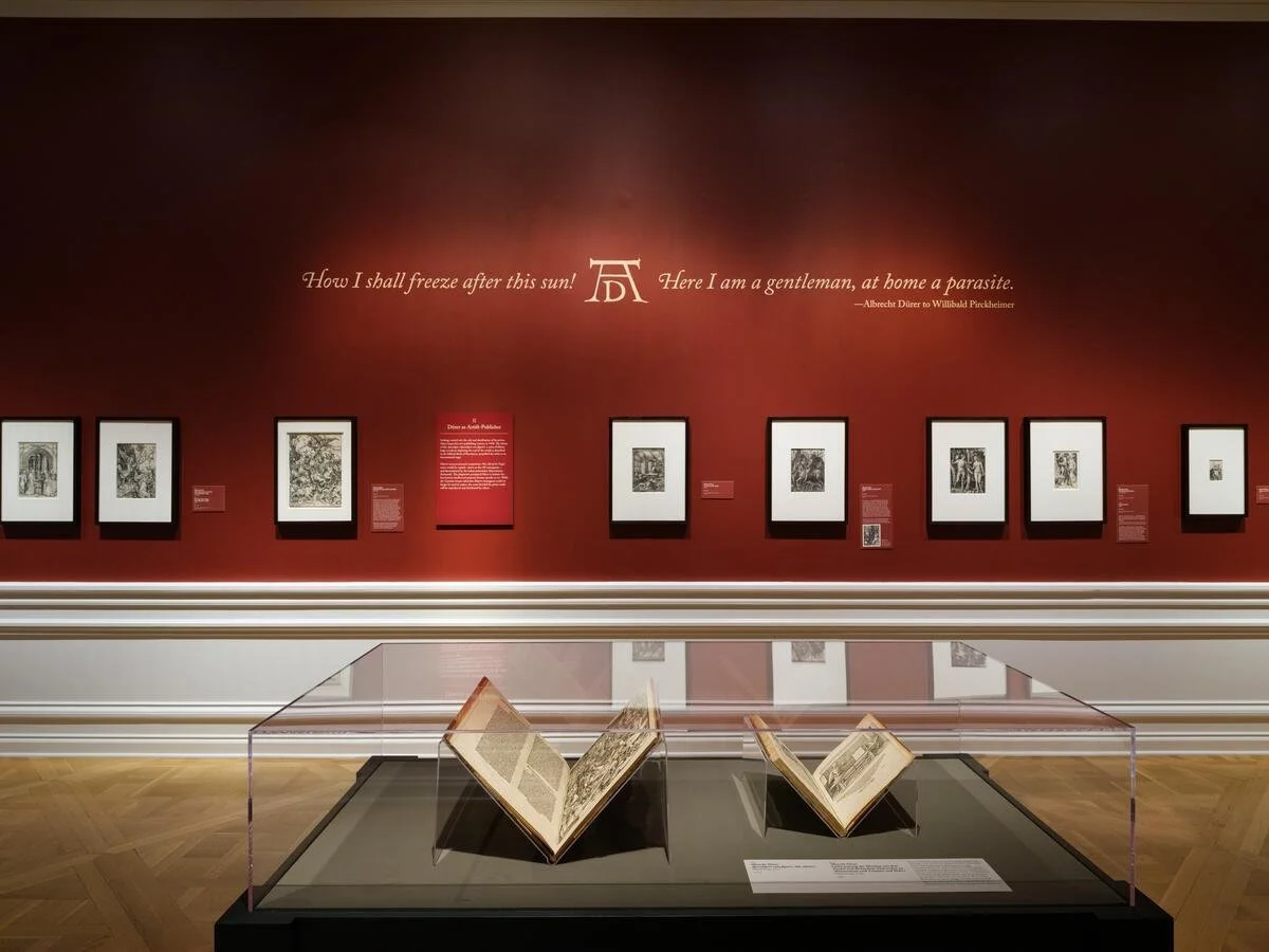 Museum exhibit featuring framed artworks on red wall and open books in a glass case with information plaques.