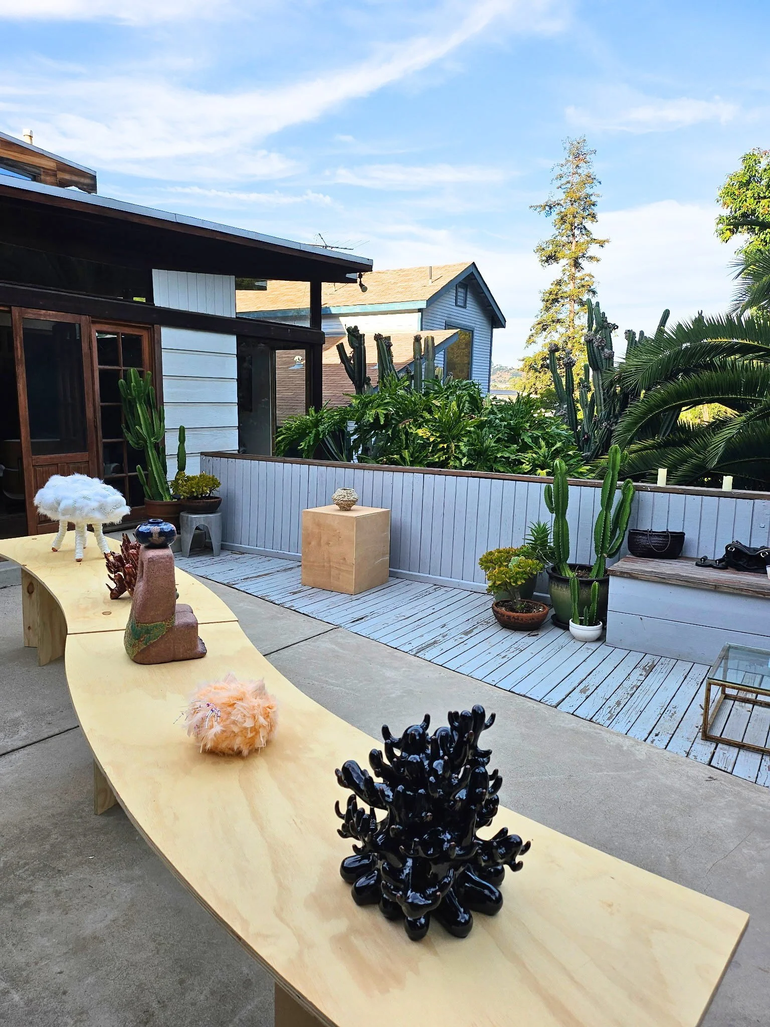 Ceramic sculptures on top of S-curve table; vase sculpture in background of outdoor patio, sky, trees, two houses