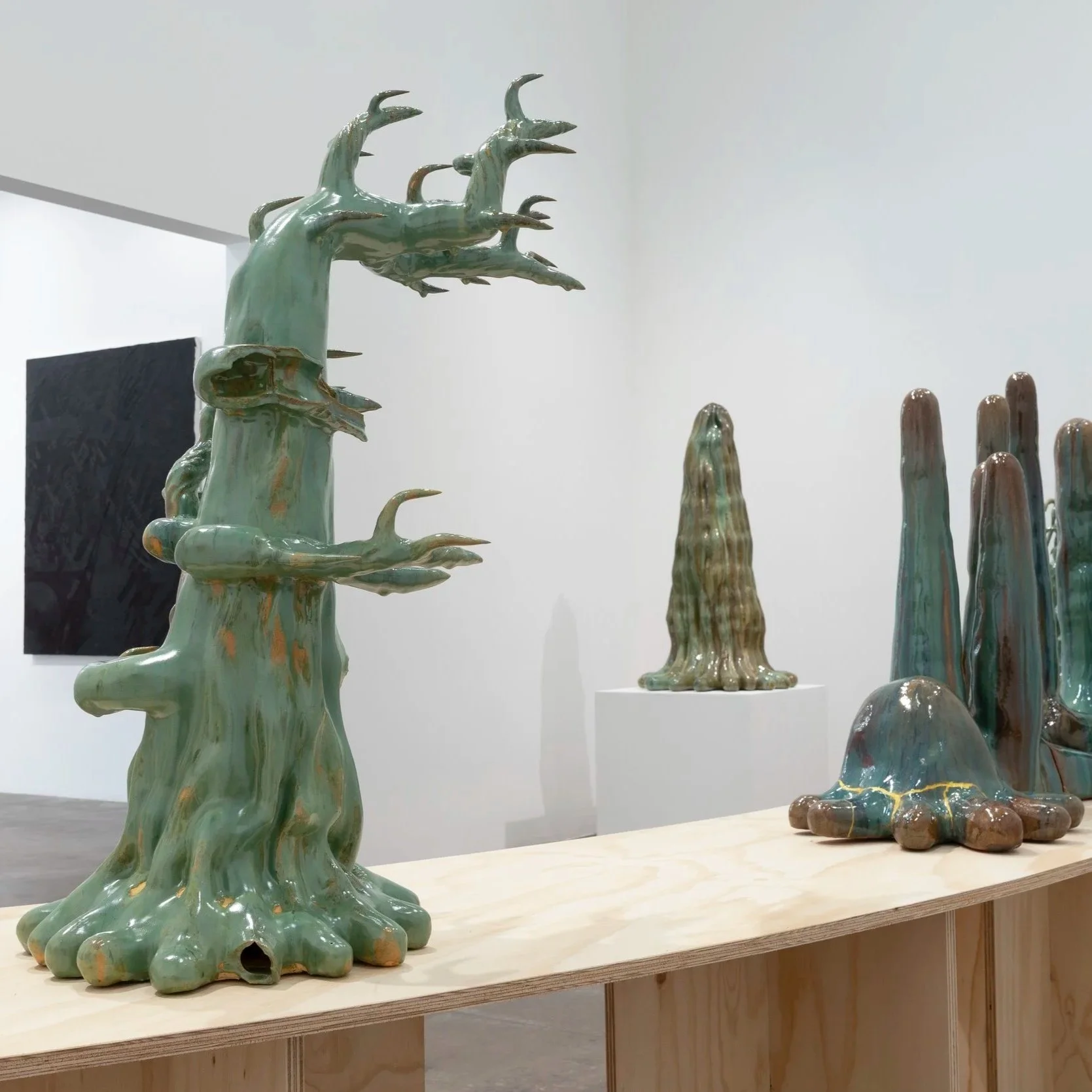 Ceramic sculptures resembling trees and organic forms displayed on a wooden table; behind on left is a black painting. All within an art gallery.