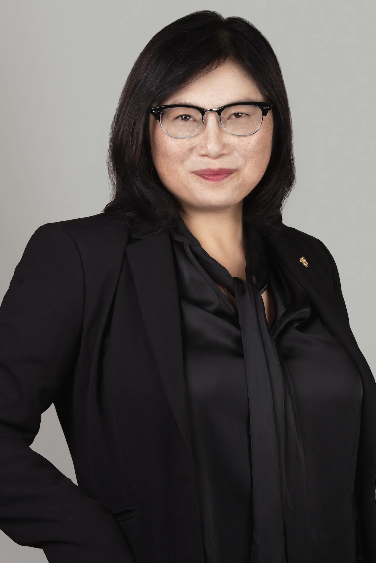 A woman with shoulder-length black hair and glasses, wearing a black blazer and blouse, standing against a plain gray background.