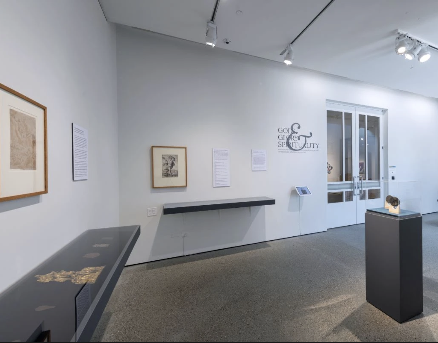 Gallery with white walls, framed drawing and framed print on the walls; round medal sculptures on pedestal, flat metal plaquettes on shelving against walls.