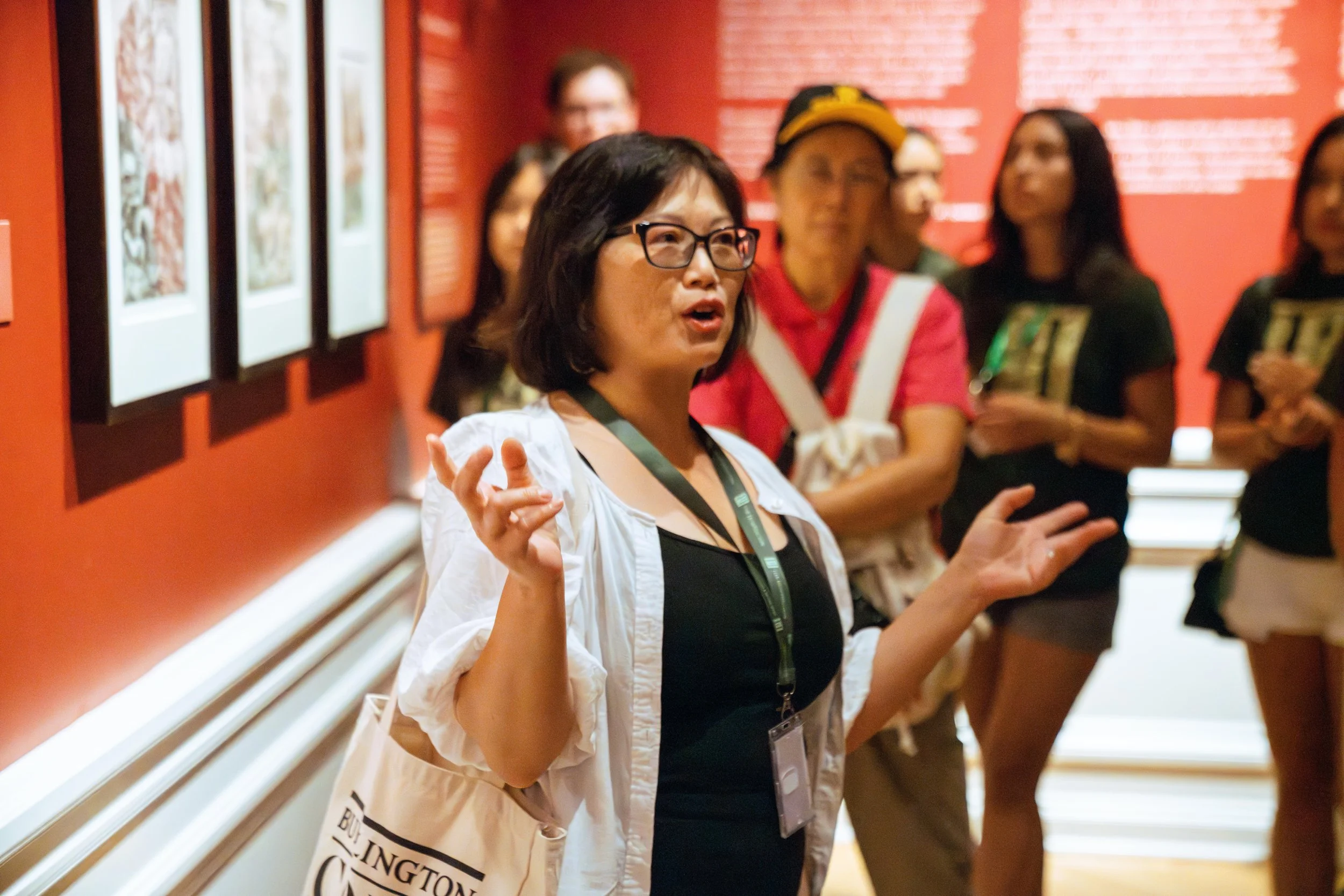 A woman wearing glasses and a white jacket speaking to a group in an art gallery, others are listening, red walls with framed artworks.