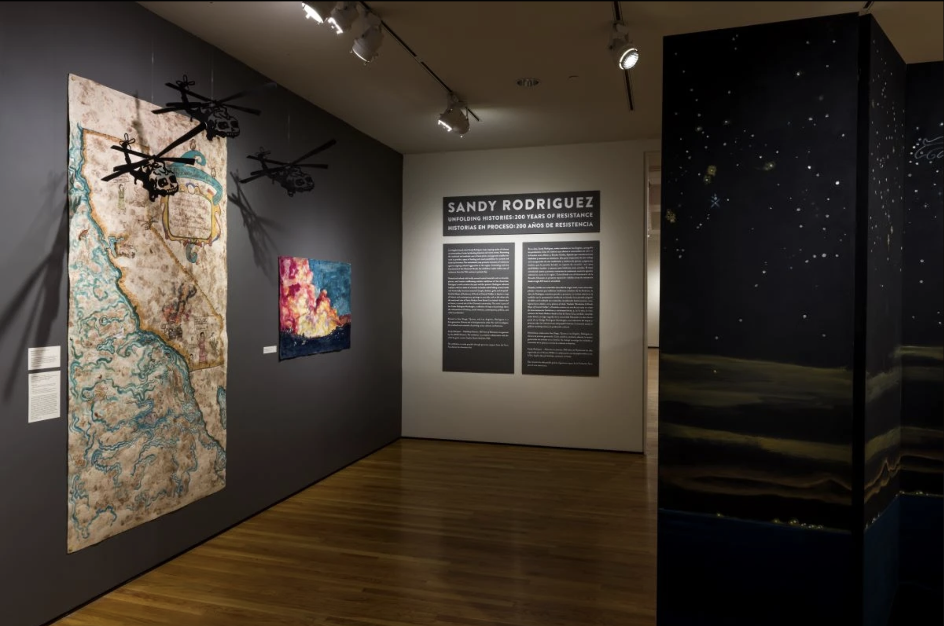 Gallery with map on left side, helicopters hanging above it, "Sandy Rodriguez / Unfolding Histories / 200 Years of Resistance" on far wall; and black upright panel with painting on right.