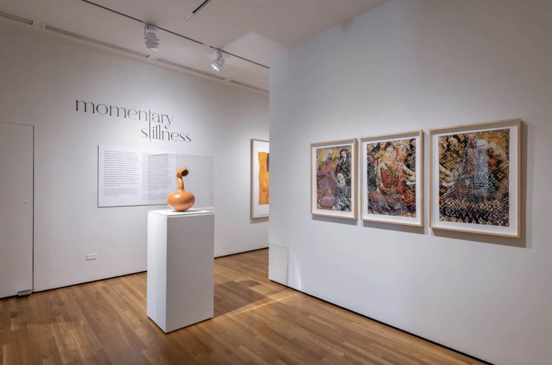 Gallery with white walls, "momentary stillness" in black font on far wall, sculpture on pedestal in middle, three framed art works on right wall.