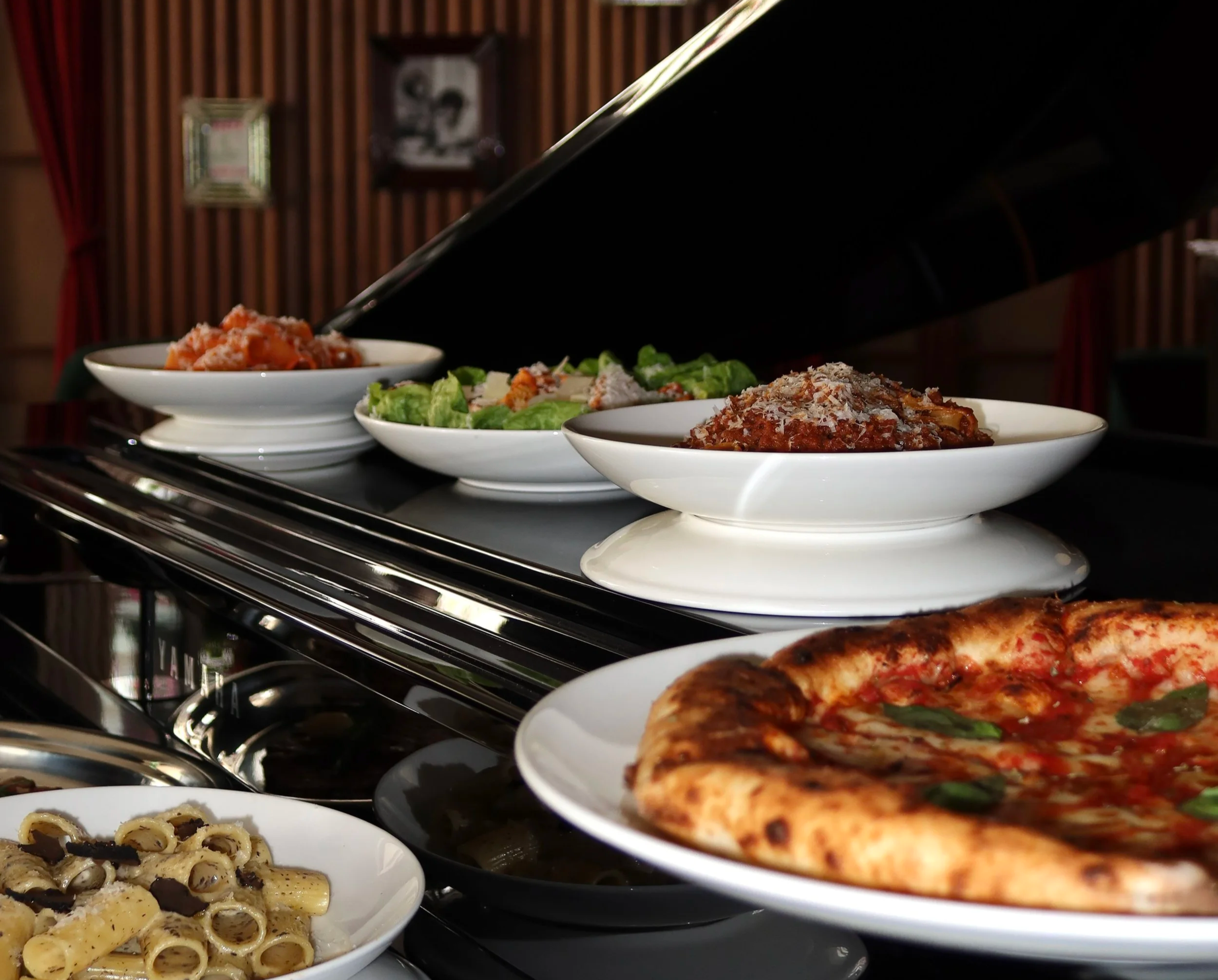 A buffet of various dishes including pasta with black truffle, salad with lettuce and shredded cheese, spaghetti bizzeria with melted cheese, and pizza with basil leaves on top.