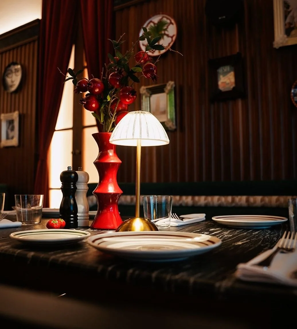 A dining table with plates, glasses, and utensils, featuring a red vase with pomegranete and leaves, and a small table lamp with a white shade set against a wooden-paneled wall with framed pictures and curtains in the background.