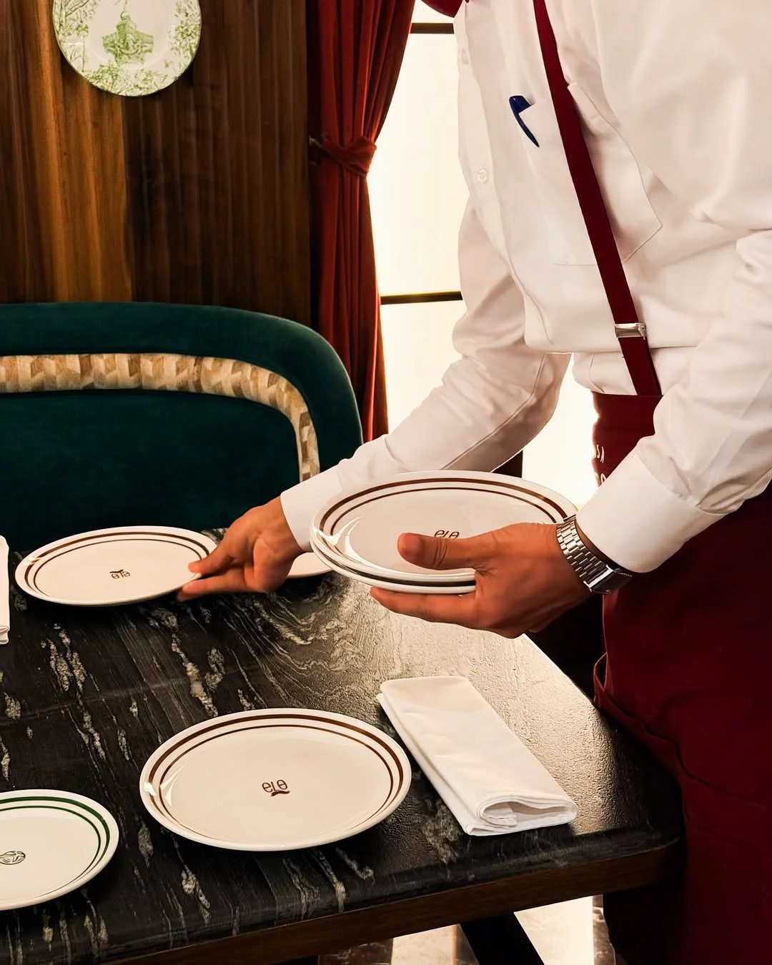 A waiter in a white shirt and red apron setting white plates with a red and green border on a dark wooden table in a restaurant or dining room.