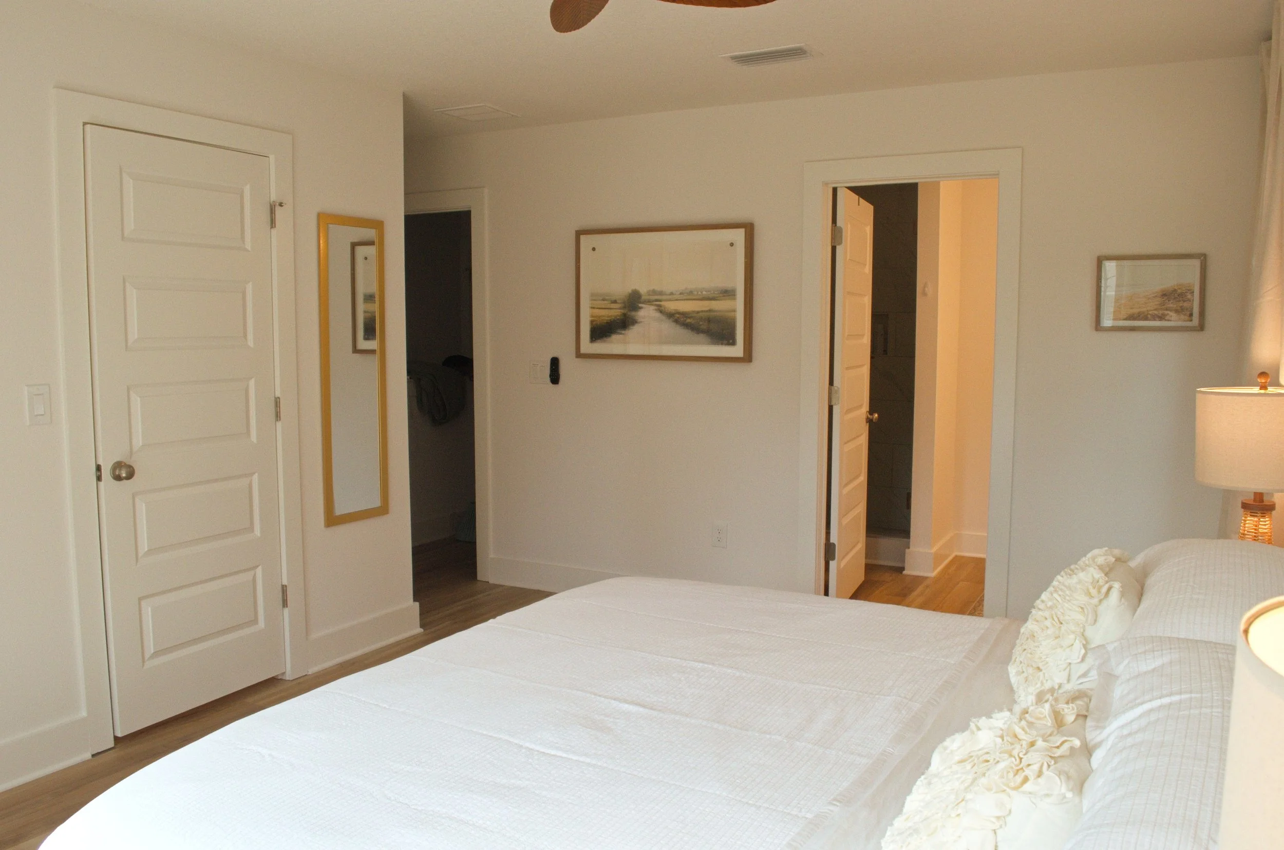 A bedroom with white walls and, white doors on the closets and room entrance.  Artwork and a mirror decorate the walls.  Partial view of a bed with white bedding and a floral pillow.  A bedside lamp on a table is visible on the right.