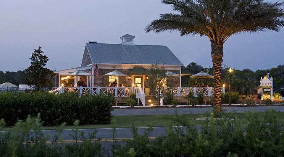 A cozy building with a porch and string lights, surrounded by greenery and a tall palm tree, during twilight.