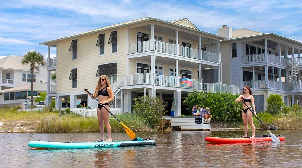 Two women paddleboarding on a calm waterway in front of a large beach house. One woman is dressed in a black swimsuit and sunglasses, holding a yellow paddle on a blue paddleboard. The other woman is also in a black swimsuit with sunglasses, holding a paddle on a red paddleboard. In the background, people are sitting outside near a green shrub, and the house has multiple balconies with amenities.